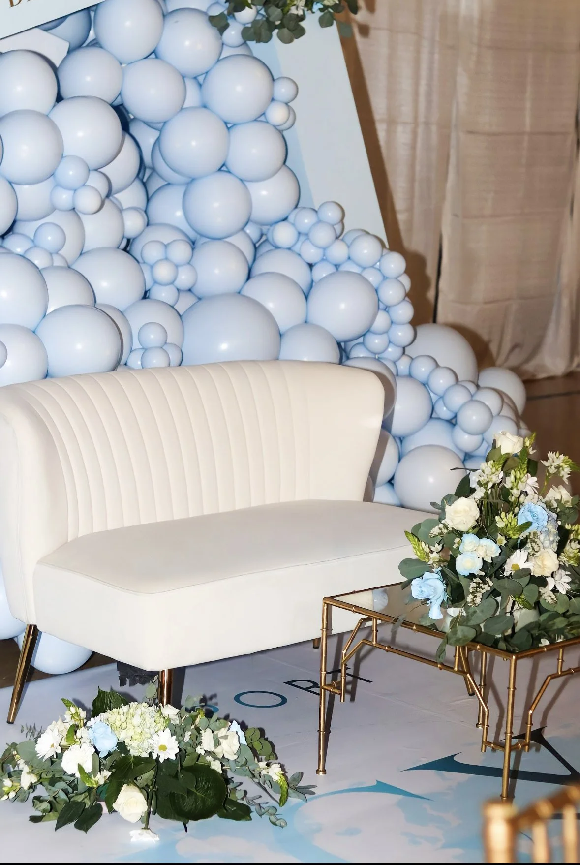 A decorative backdrop made of white and light blue balloons with a white velvet sofa and a gold metal table with a floral arrangement of white and blue flowers and greenery in front of it.