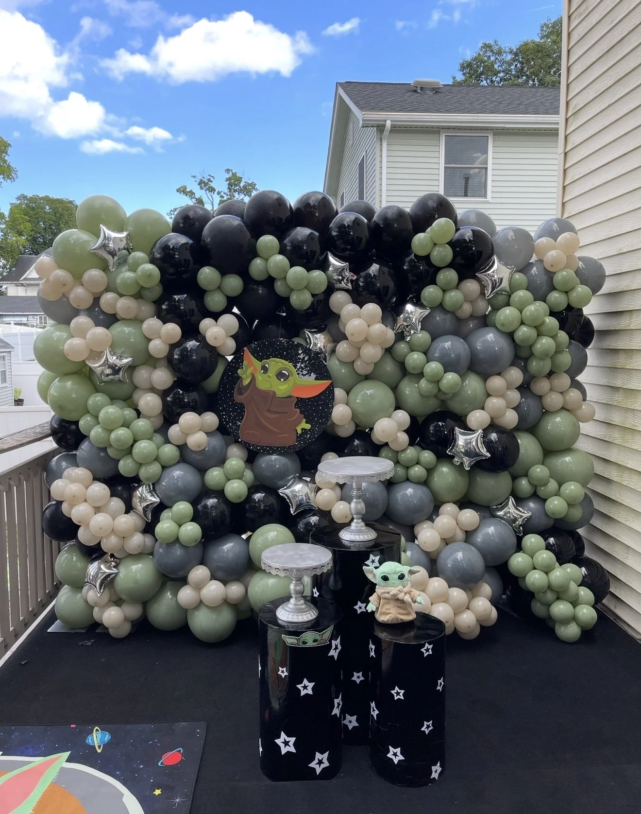 Decorative balloons arranged in a space-themed galaxy pattern with green, black, cream, and gray balloons, silver star-shaped balloons, and a sign featuring Baby Yoda from Star Wars. There are small tables with starry designs and Baby Yoda plush on a black tablecloth, set up on a porch with a house and blue sky with clouds in the background.