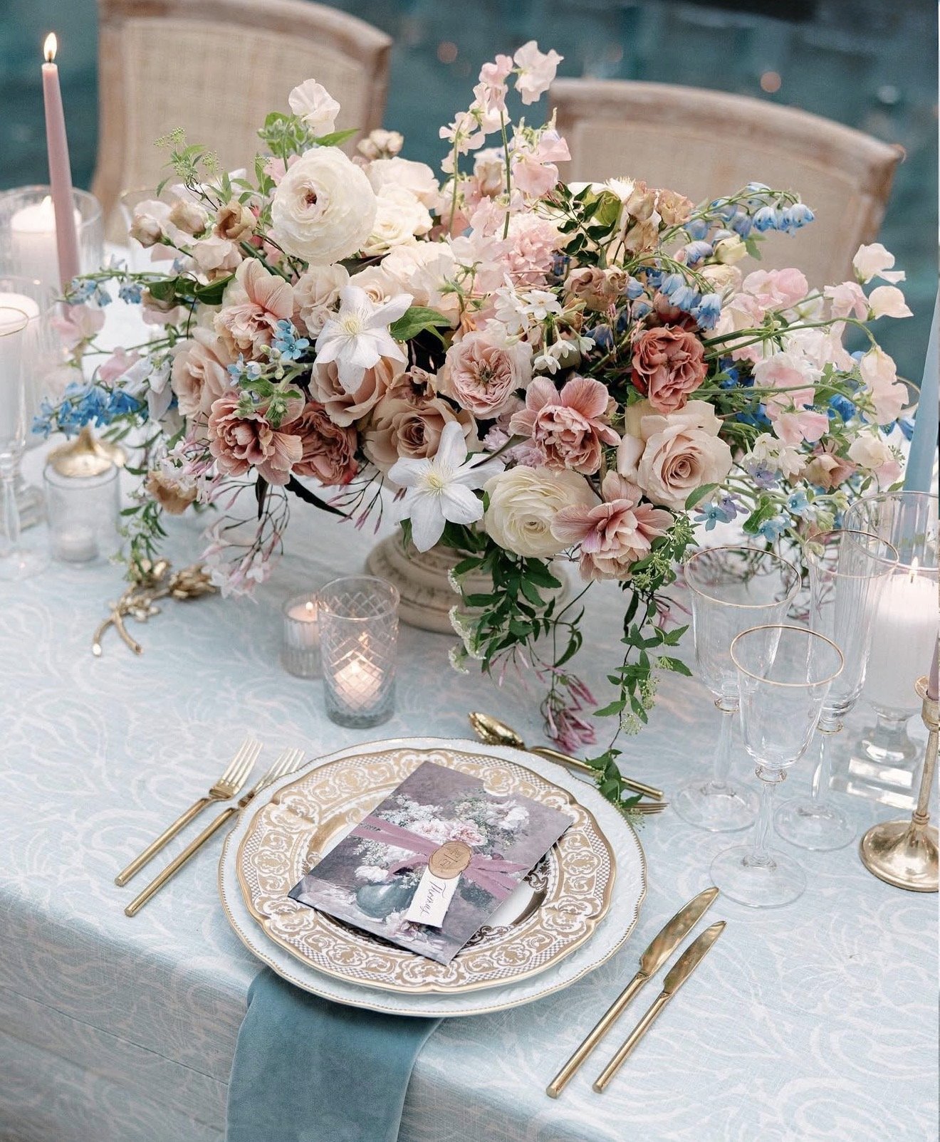Elegant table setting with a large pastel pink and white floral centerpiece, gold flatware, intricately patterned gold-edged plates, clear glassware, and lit candles on a light blue tablecloth for a formal event.