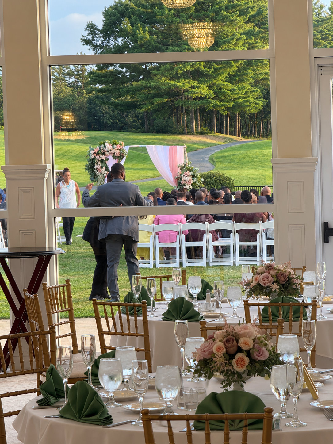 View from inside a decorated banquet hall looking out a window at an outdoor wedding ceremony setup on a grassy field, with chairs and floral arrangements.