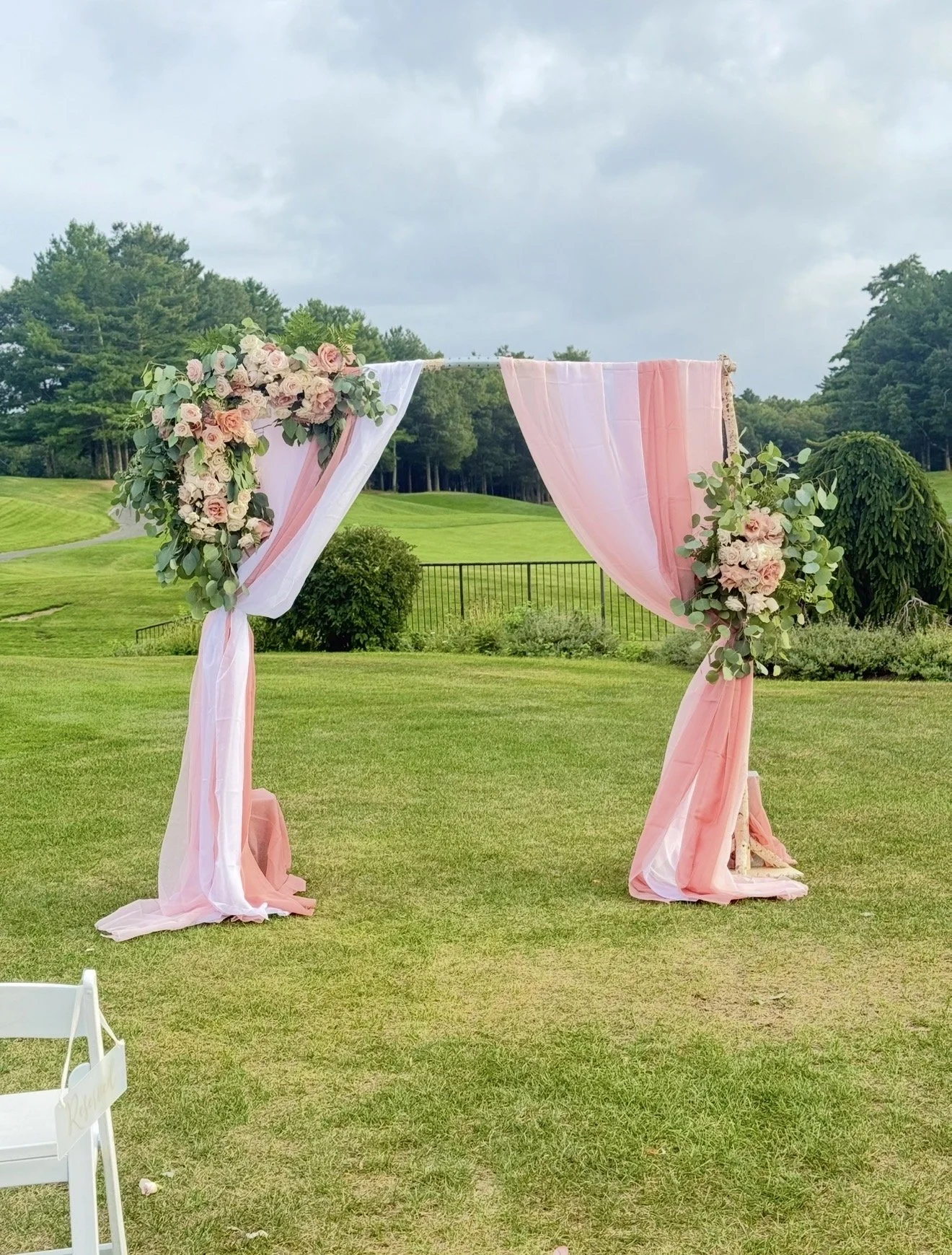 A pink and white floral wedding arch on a grassy outdoor lawn, with trees and cloudy sky in the background.