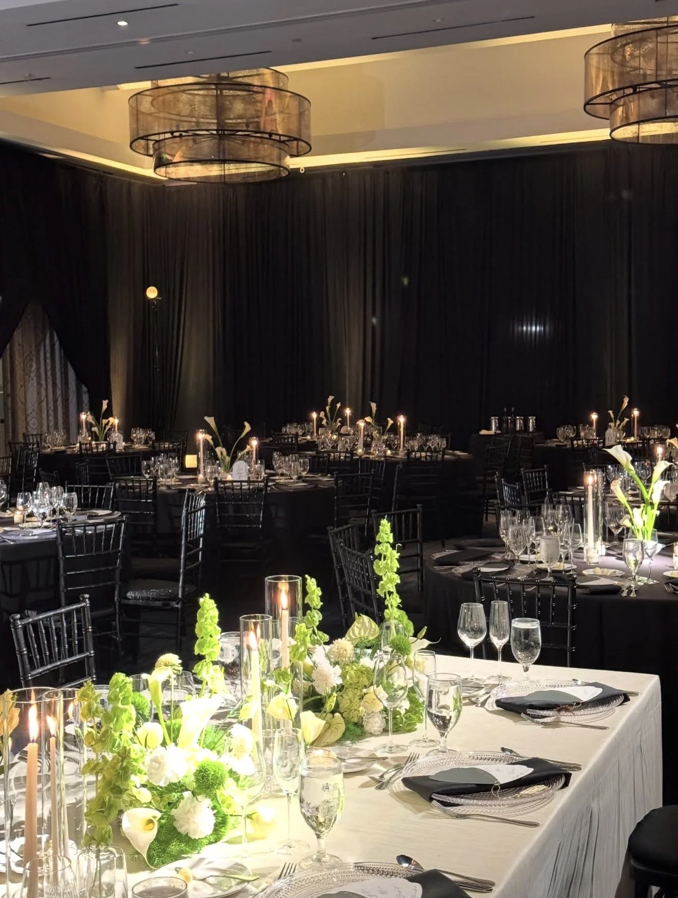 Elegant banquet hall decorated for a formal event, with round tables, candlelit floral centerpieces, and black curtains in the background.