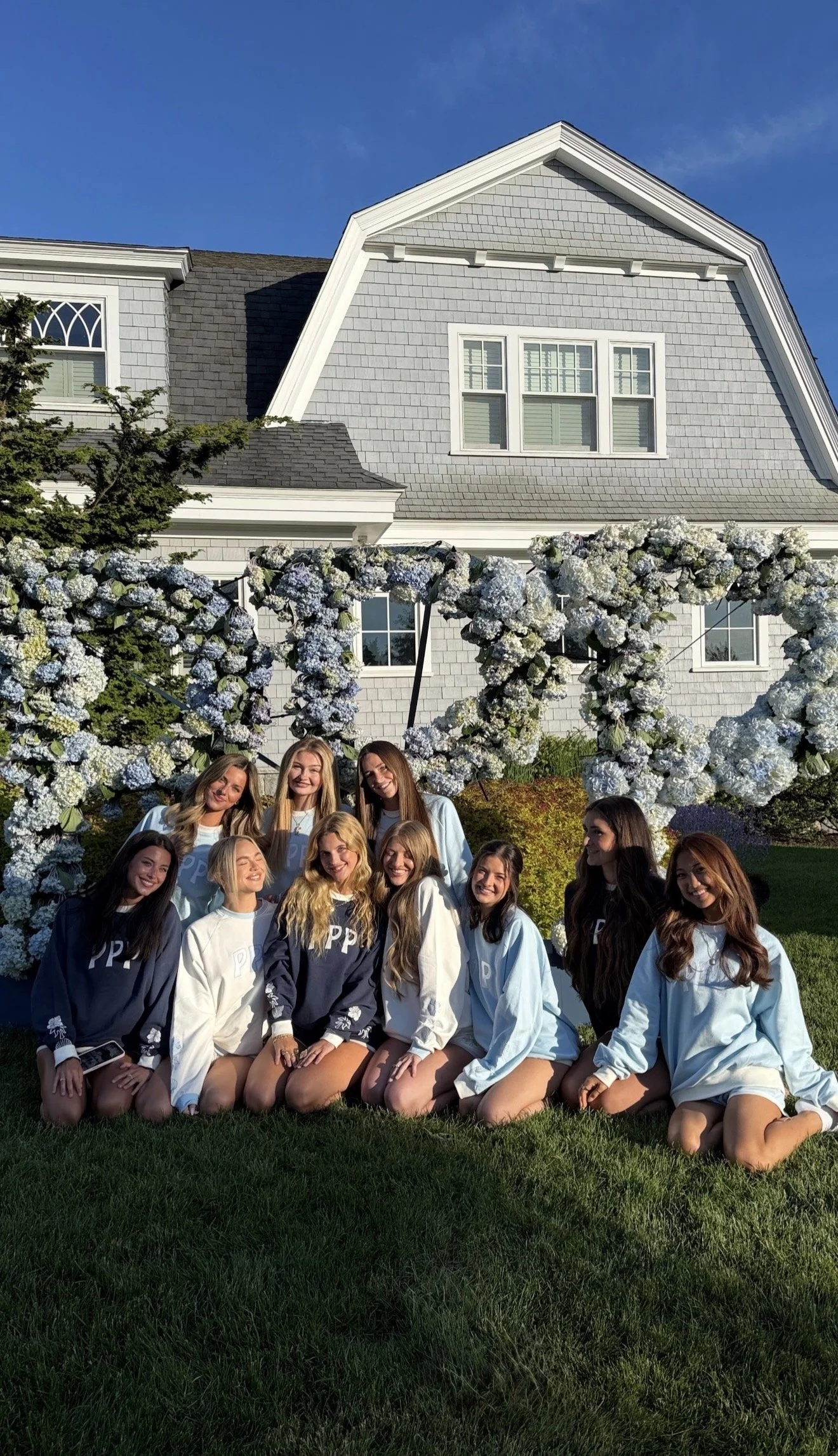 A group of young women in pajamas, kneeling and sitting on grass in front of a floral arch at an outdoor event on a sunny day.