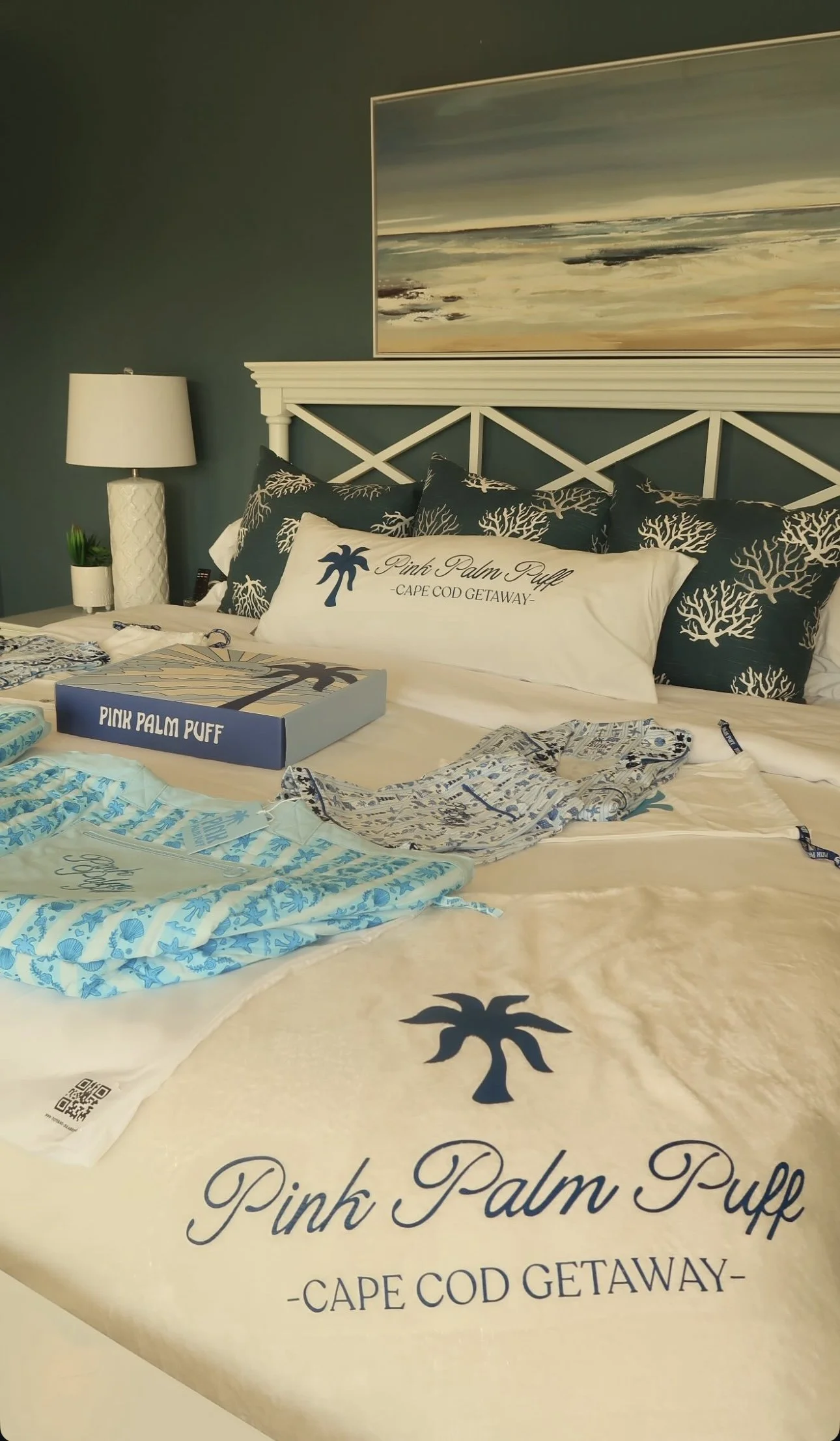 A coastal hotel bed with pillows featuring navy and white coral patterns, a white pillow with a palm tree and text, and a duvet with blue palm tree designs. A framed landscape painting of the beach hangs above the bed. A white lamp and small plant are on a nightstand.