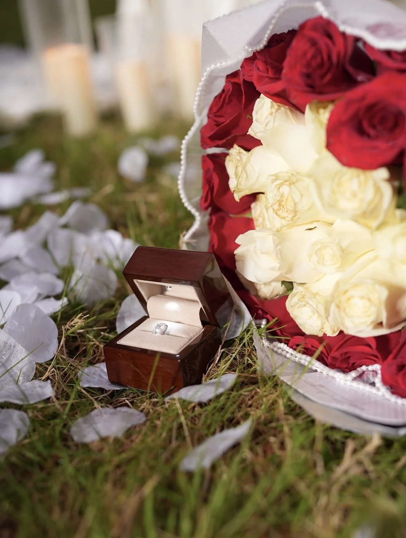 A bouquet of red and white roses, a jewelry box with an engagement ring, candlelights, and scattered white petals on grass, suggesting a romantic proposal setting.