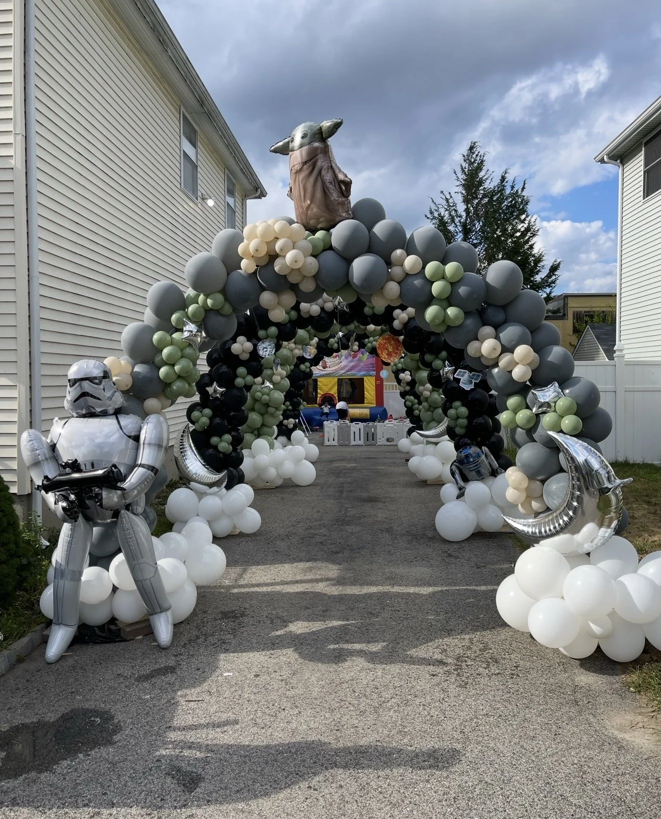 A Star Wars-themed balloon archway decorated with black, white, gray, and green balloons, featuring a large Baby Yoda (Grogu) figure at the top and Star Wars character figures made of balloons at the bottom, with a bounce house in the background.