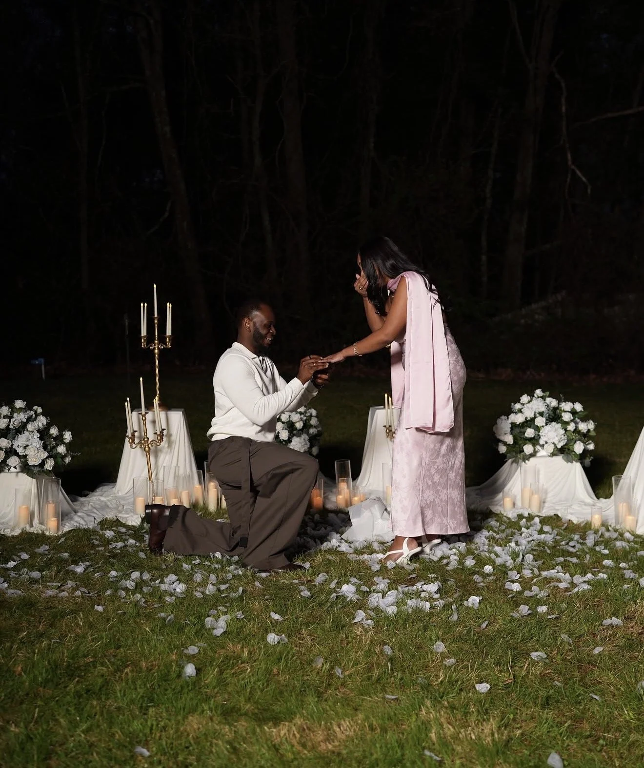 A man proposing to a woman during an outdoor nighttime romantic setting, surrounded by candles and white floral decorations.