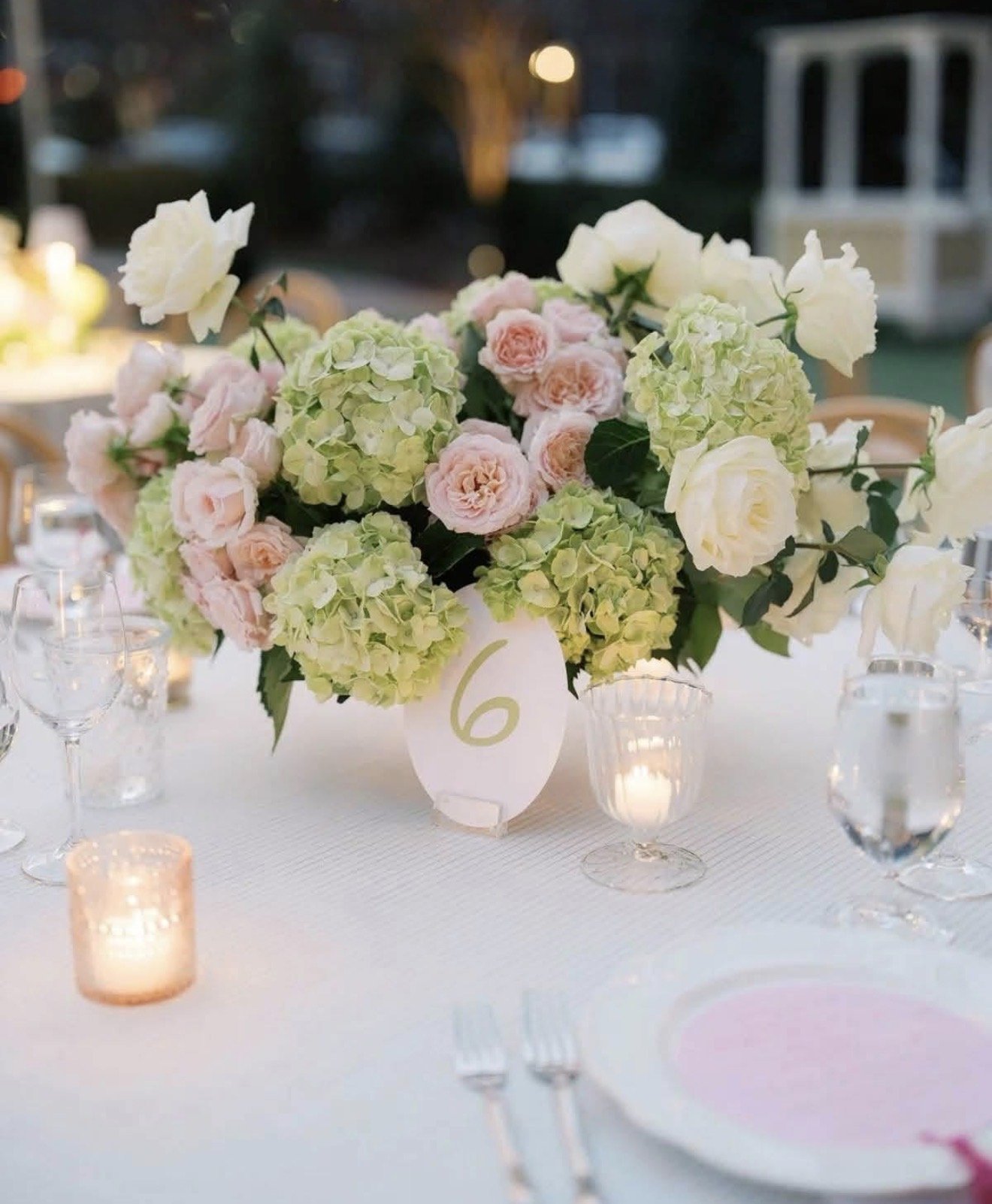 Elegant table centerpiece with white and pale pink roses and green hydrangeas, table number 6 card, surrounded by lit candles, wine glasses, and tableware at an outdoor evening event.