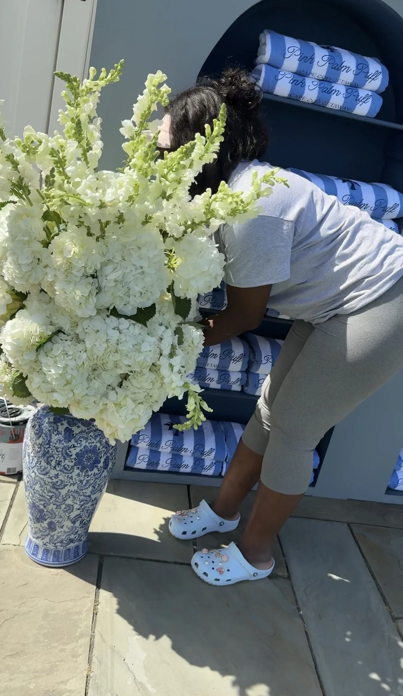 A woman bending over in front of a large bouquet of white flowers in a decorative blue and white vase, with a stack of blue and white towels or blankets labeled "Pink Palm Puff" in the background.