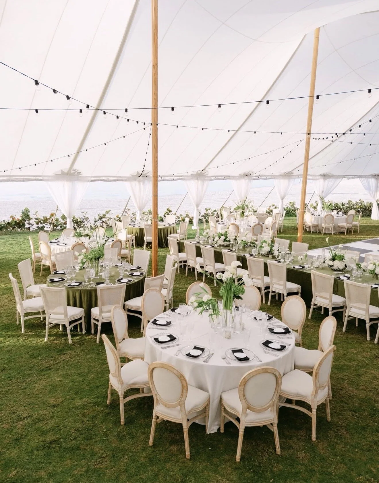 Wedding reception setup with round tables covered with white linens, black and white place settings, and floral centerpieces, under a white tent with string lights, overlooking a body of water.