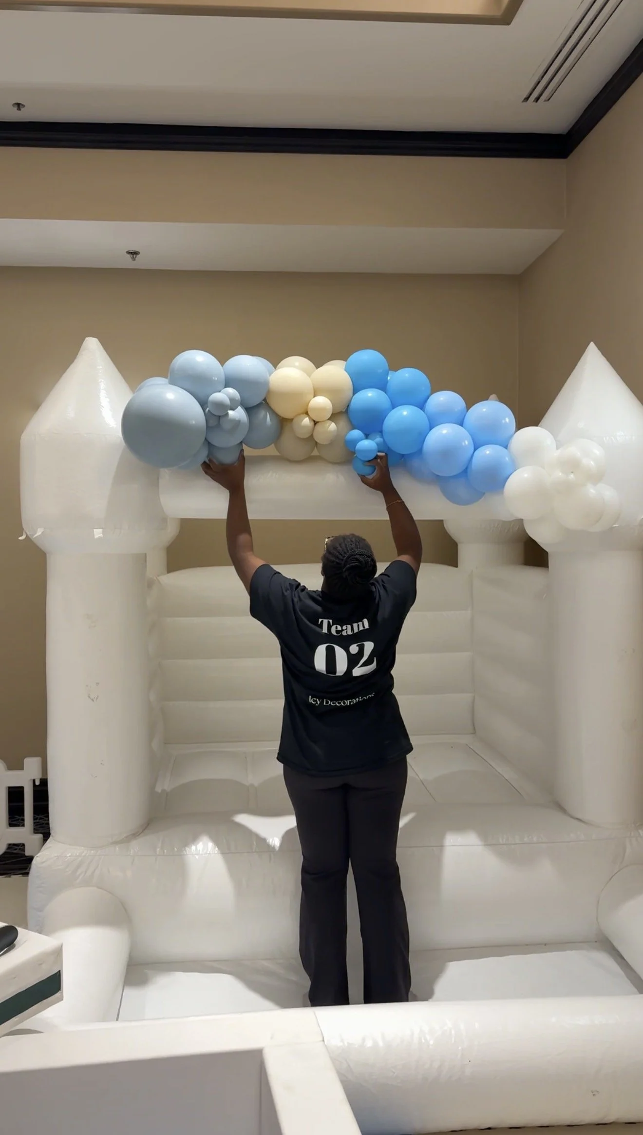 Person decorating a large inflatable castle with blue, cream, and white balloons inside a room.