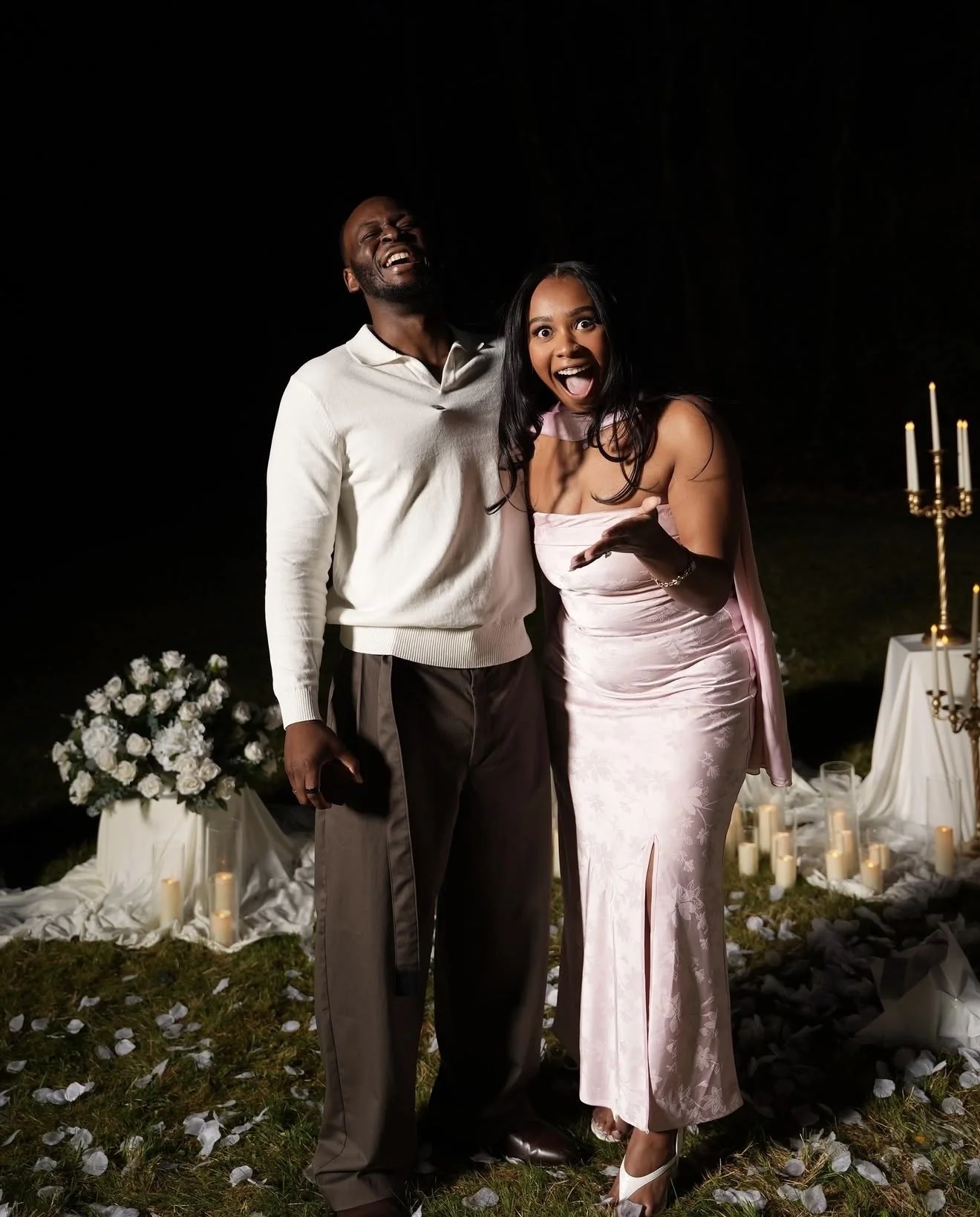 Two people, a man and a woman, smiling and celebrating outdoors at night, standing on grass with scattered flower petals, candles, and white floral arrangements and candles in the background.