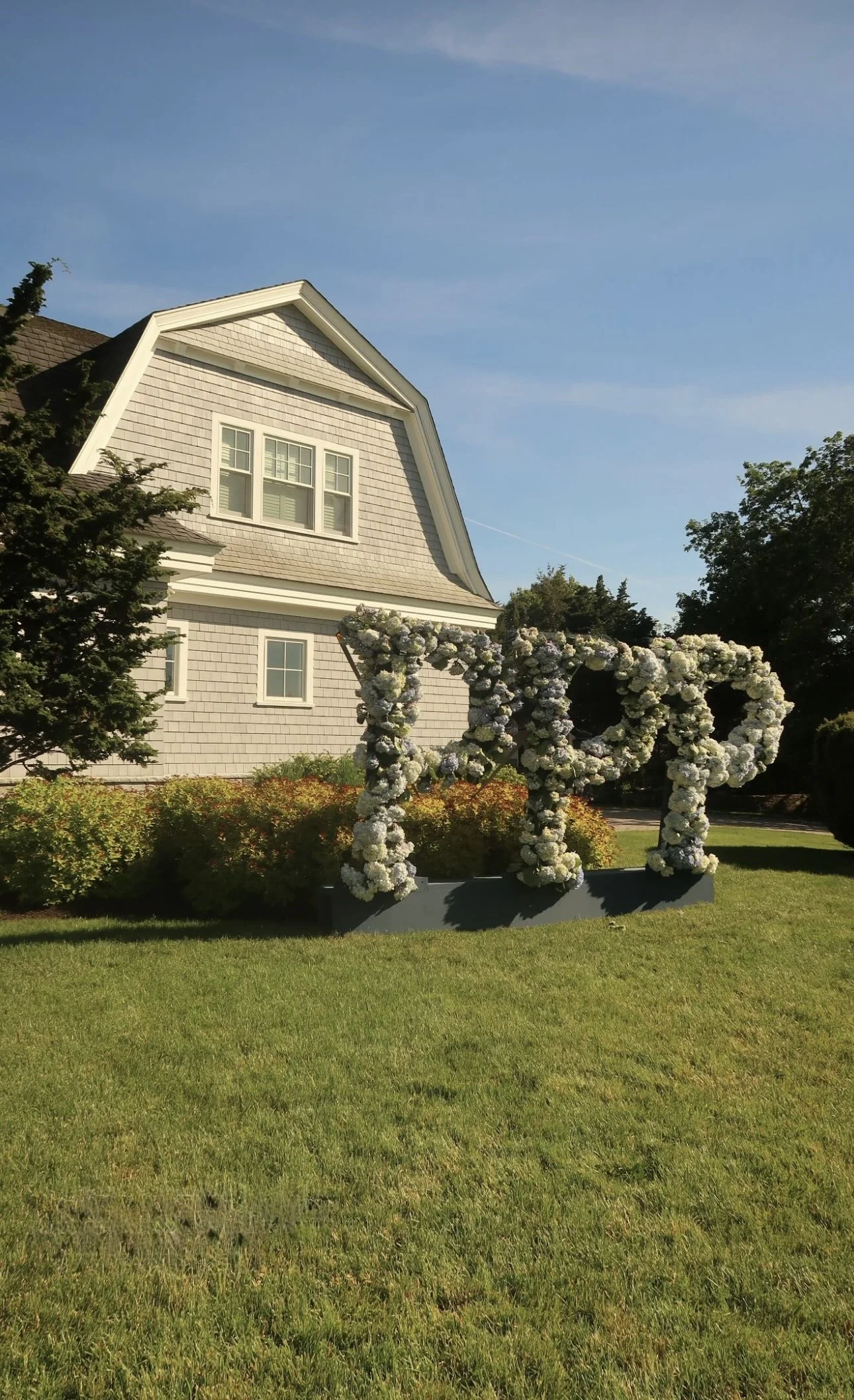 A house with beige shingles and white trim under a clear blue sky, surrounded by green trees and a lawn, with a floral art installation spelling 'LOVE' in front.