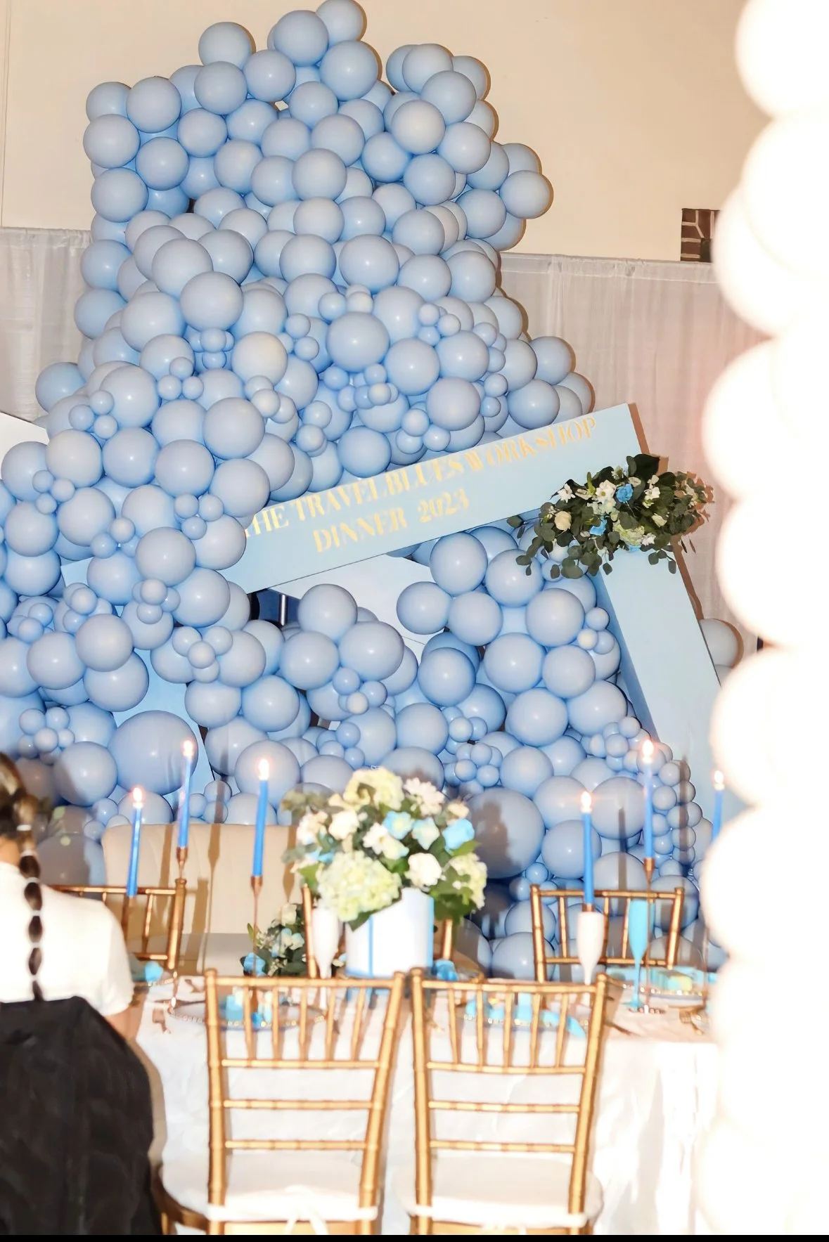 Balloon arrangement with a sign that says 'The travel boutique at Dior Shop Danner 2023' at an event decorated with flowers and candles.