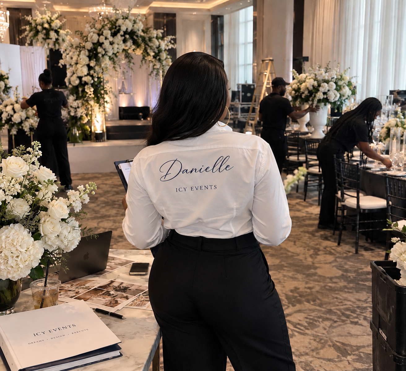 An event staff member standing at a table with floral arrangements, viewing a tablet in a decorated banquet hall with white flowers and elegant table settings.