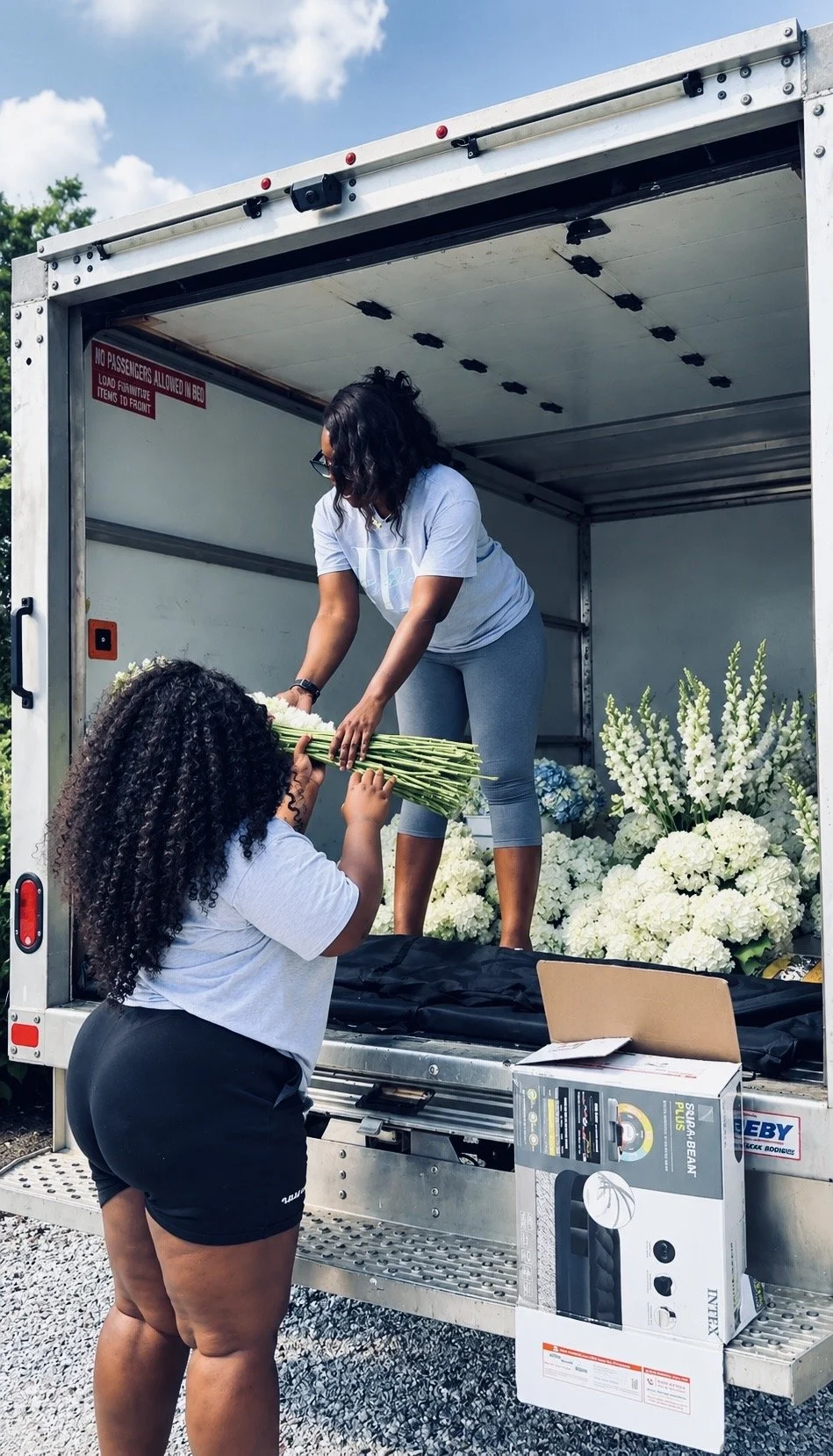 Two women unloading white flowers from a truck, with one woman on the truck handing flowers to the woman standing outside.