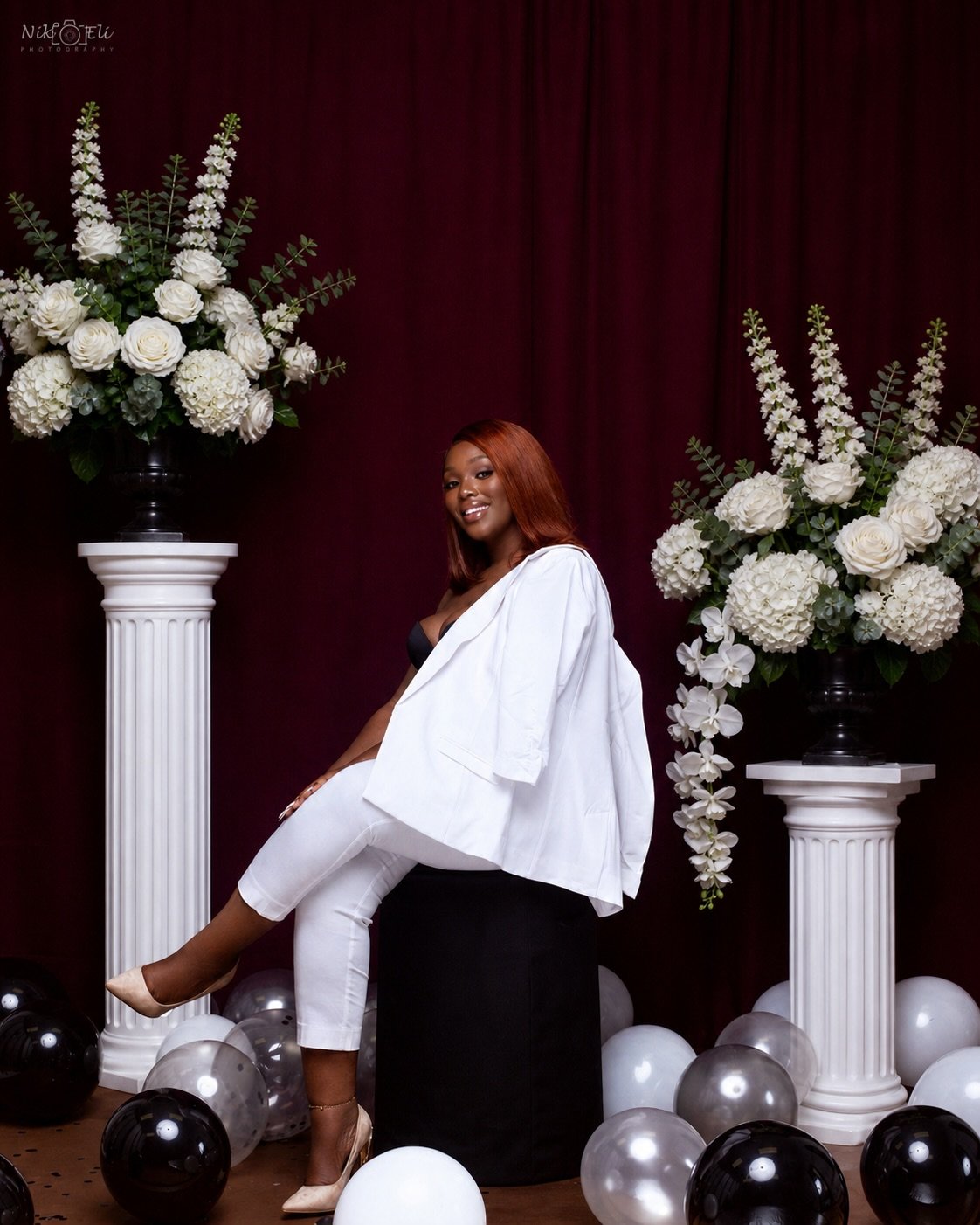 A woman with shoulder-length red hair sitting on a black stool, dressed in white pants, a black top, and a white blazer, surrounded by large white and black balloons and two tall white floral arrangements with white roses, hydrangeas, and other flowers against a burgundy curtain background.