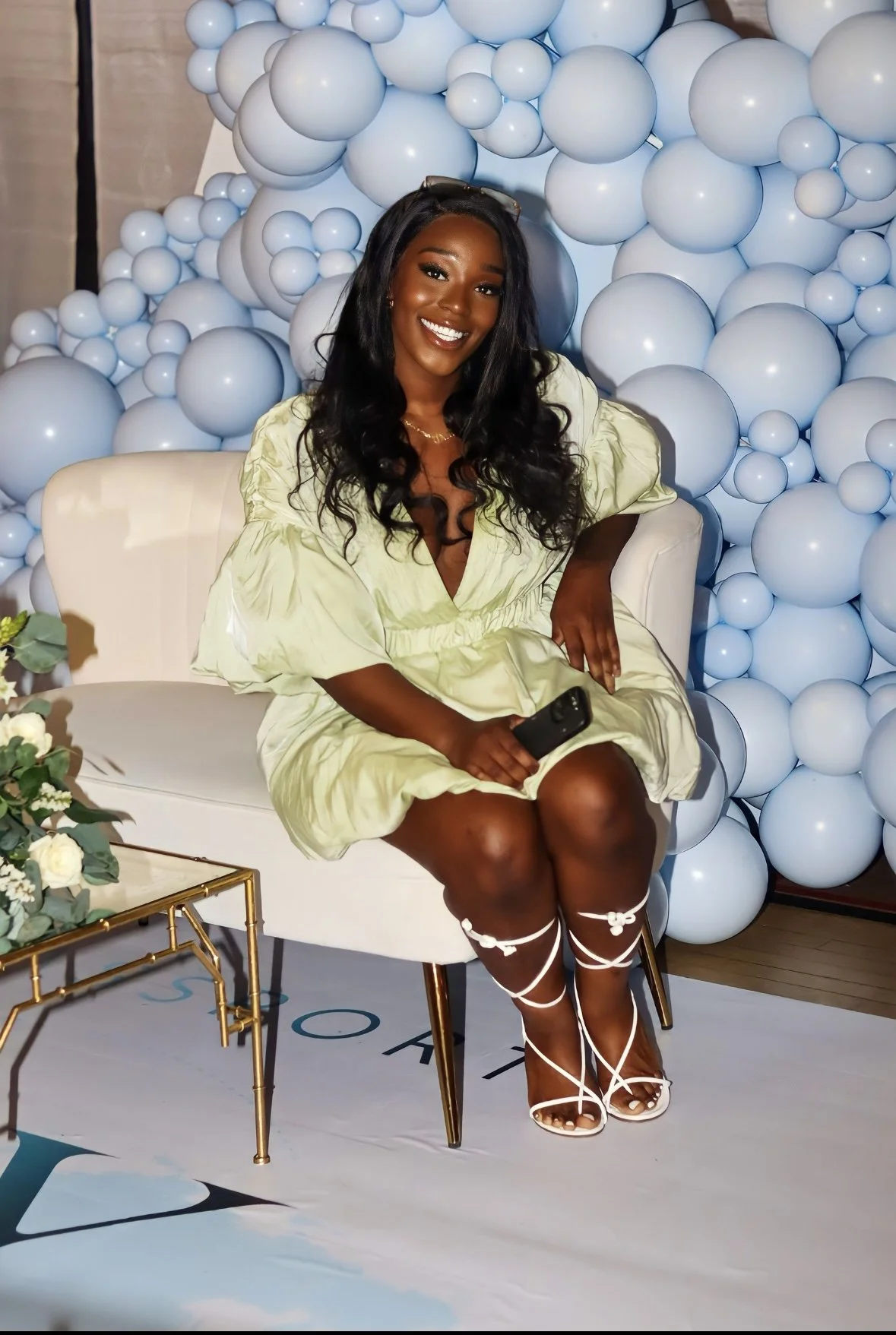 A woman with dark hair, smiling, sitting on a white chair in front of a wall decorated with white balloons, holding a black phone, wearing a light yellow dress and white strappy high-heeled sandals.