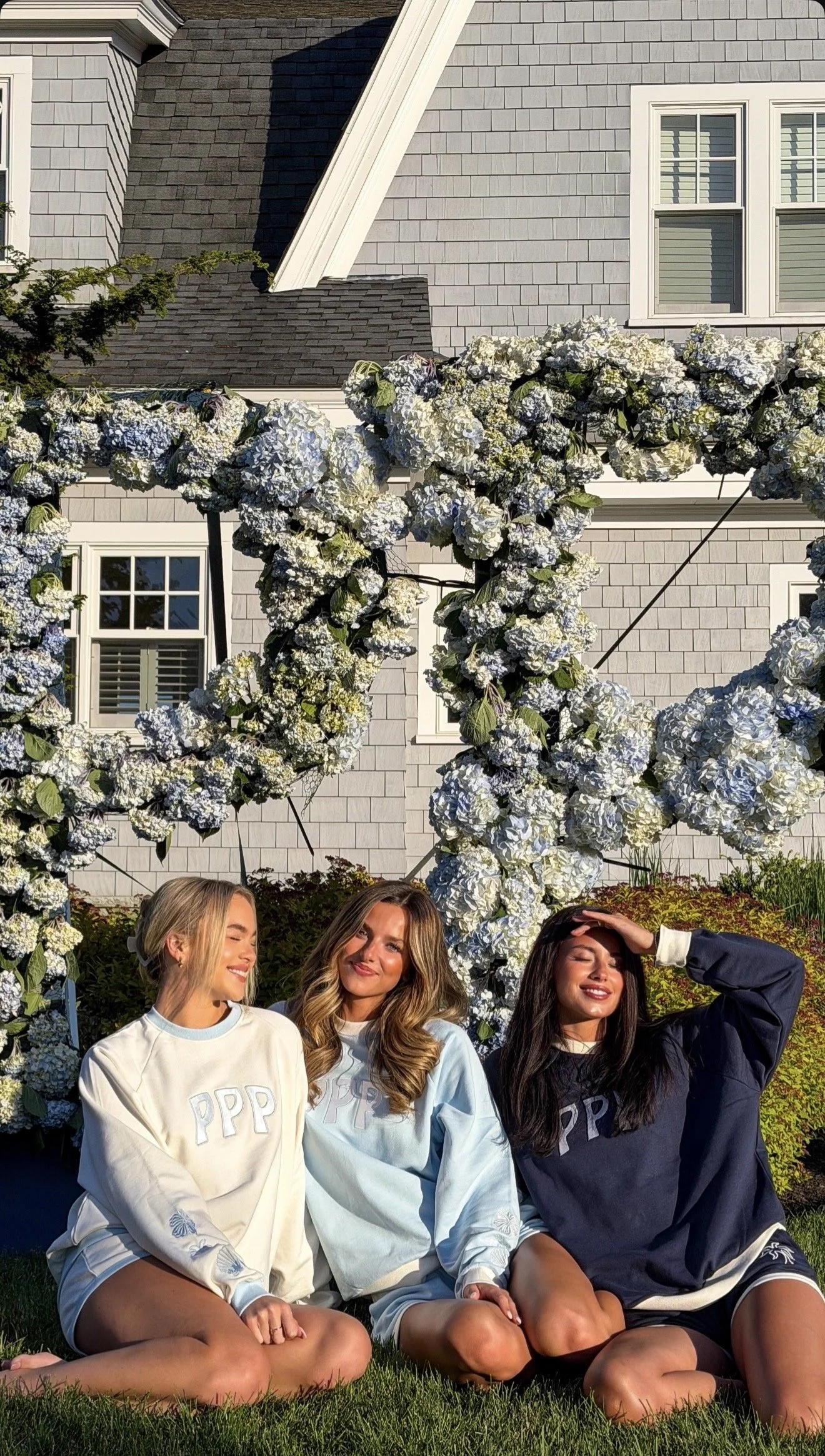 Three young women sitting on grass in front of a floral arch with a house in the background. The women are smiling and wearing sweatshirts with 'PPP' on them, during a sunny day.
