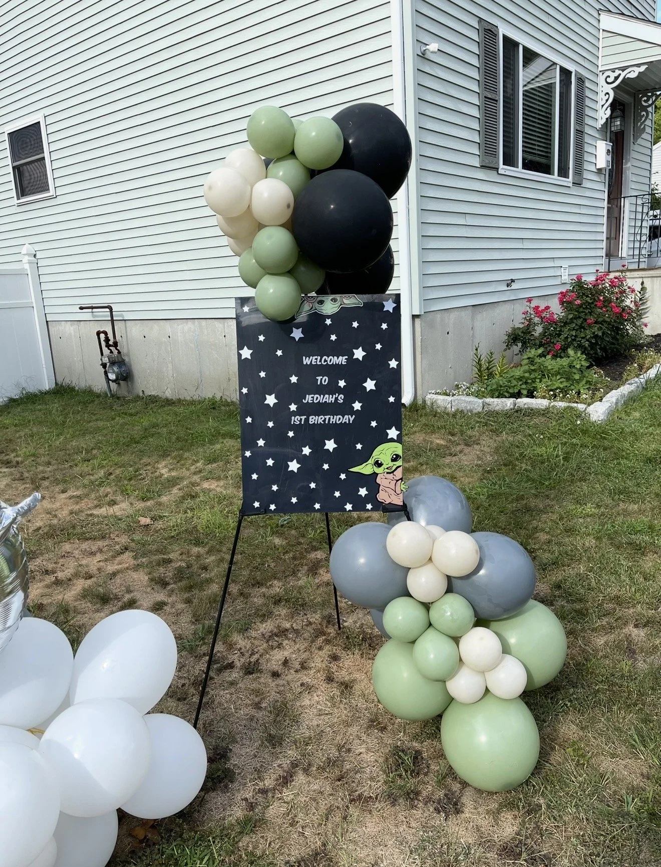 Front yard birthday party setup with black, white, and pastel green and gray balloons, and a sign with Star Wars and Baby Yoda themes reading 'Welcome to Jediah's 1st Birthday' next to a house with gray siding, a flower bed with pink blossoms, and balloon arrangements on the grass.