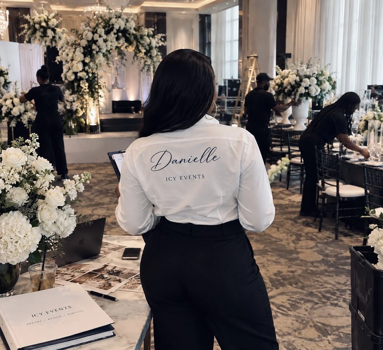 A woman with a shirt that says "Danielle ICY EVENTS" is standing in a decorated event venue with floral arrangements, a stage, and staff preparing the venue for a formal event or wedding.