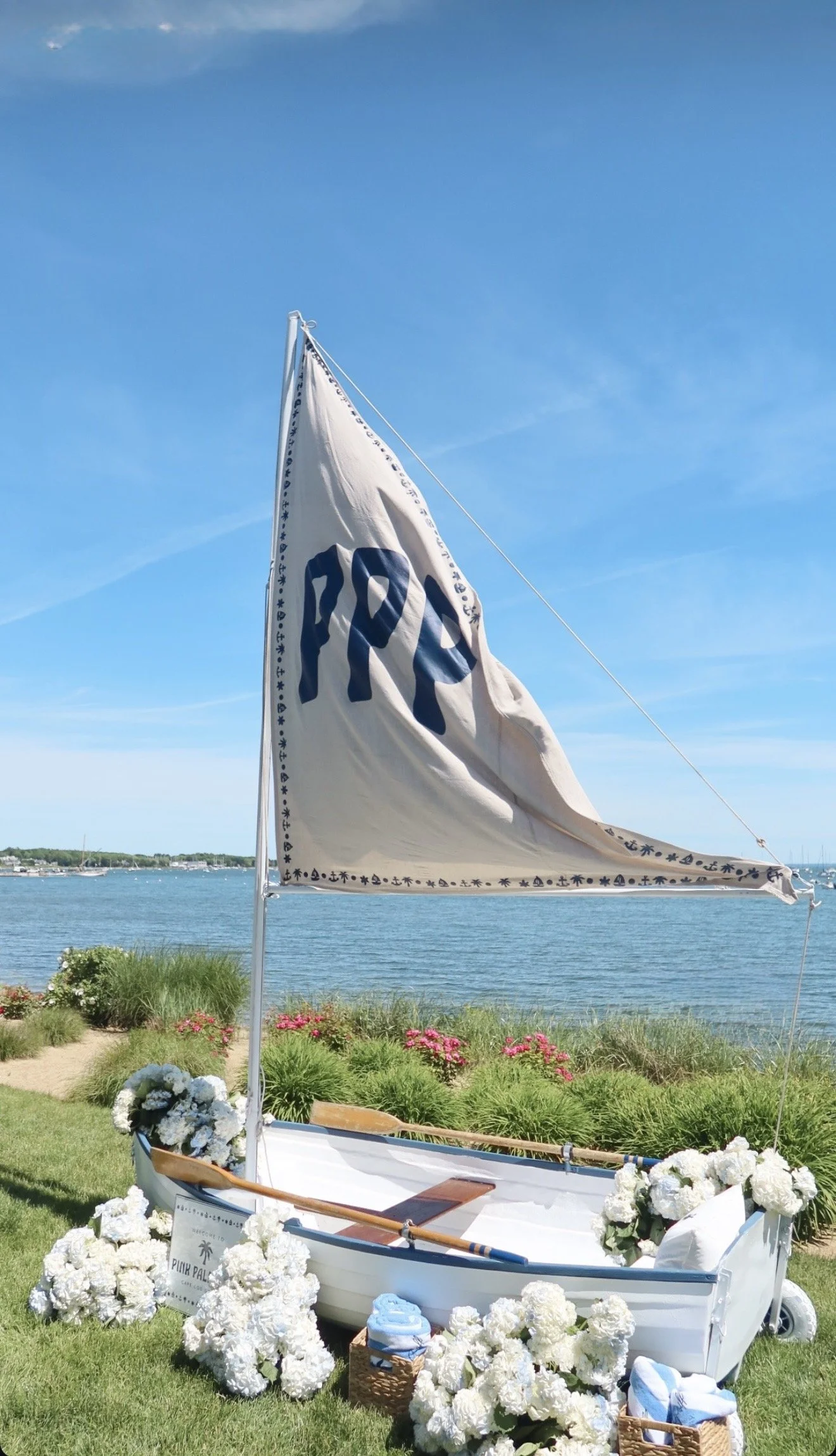 A small white boat decorated with white flowers, with a sail that has the logo 'PON', is placed on a grassy area by a body of water. The background shows a blue sky with some clouds and a distant shoreline with trees and boats.