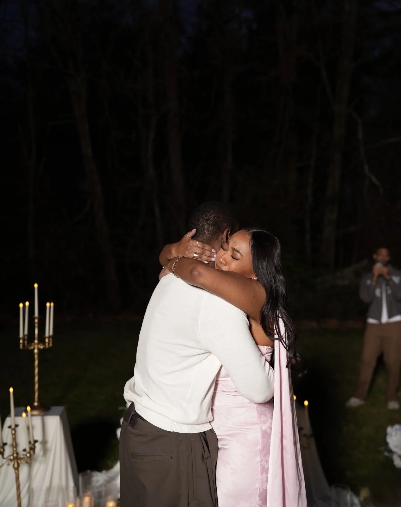 A couple sharing a heartfelt embrace at an outdoor event at night, with a woman in a pink dress and jewelry hugging a man in a white sweater, candles on tables, and a photographer capturing the moment in the background.