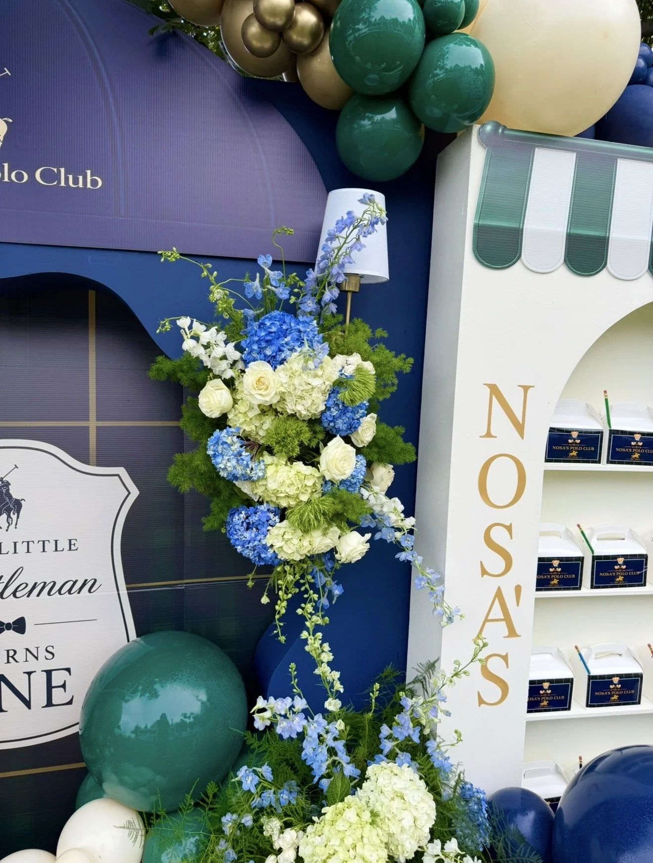 Decorative display with green, cream, and blue balloons, a large floral arrangement of white roses, blue hydrangeas, and green foliage, and a white stand with gold lettering reading 'NOSA'S'. A blue sign with white text is partially visible, featurin