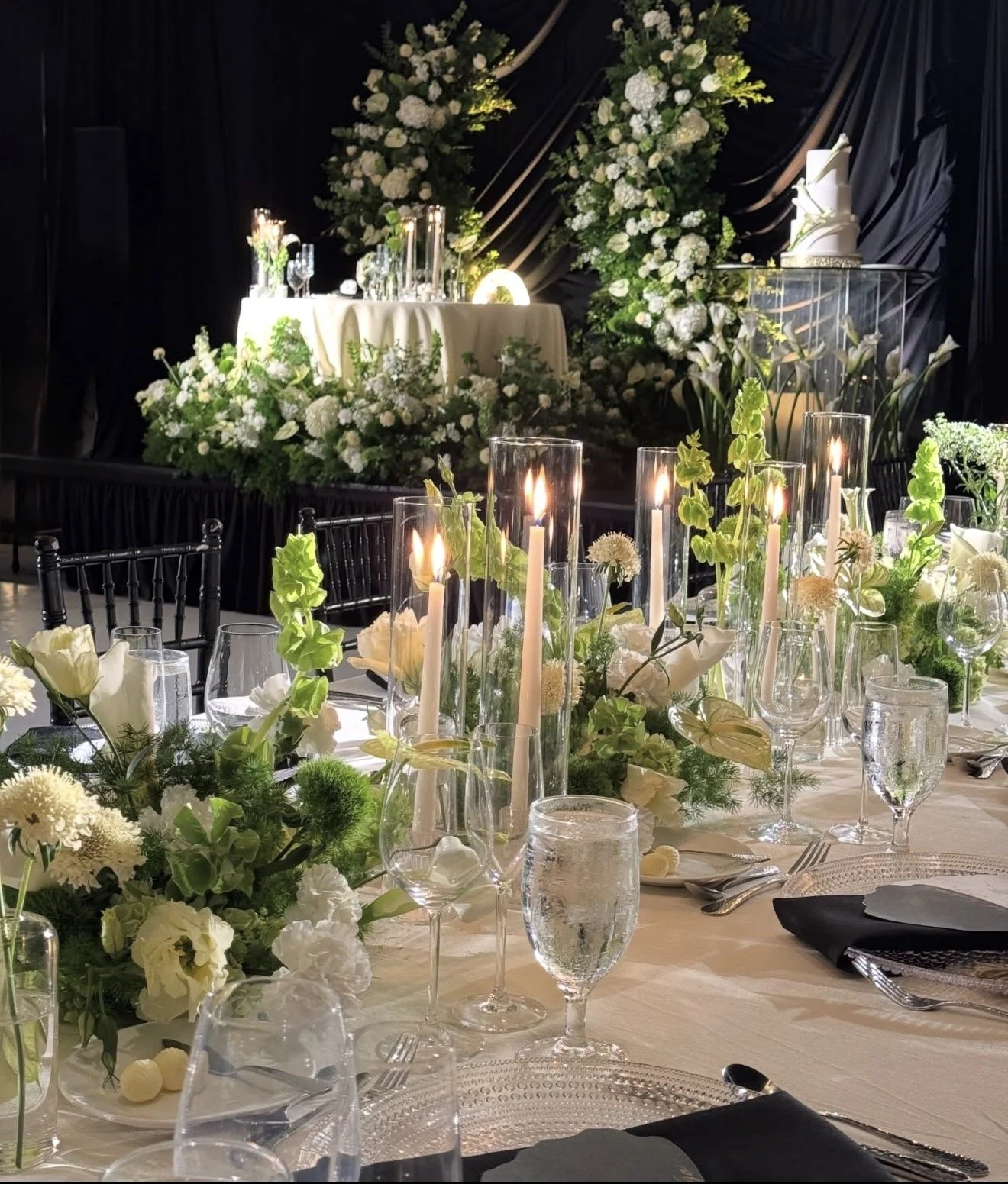 Elegant wedding or event reception decorated with white flowers, tall candles, and glassware on a table, with a floral arch and tiered cake in the background.