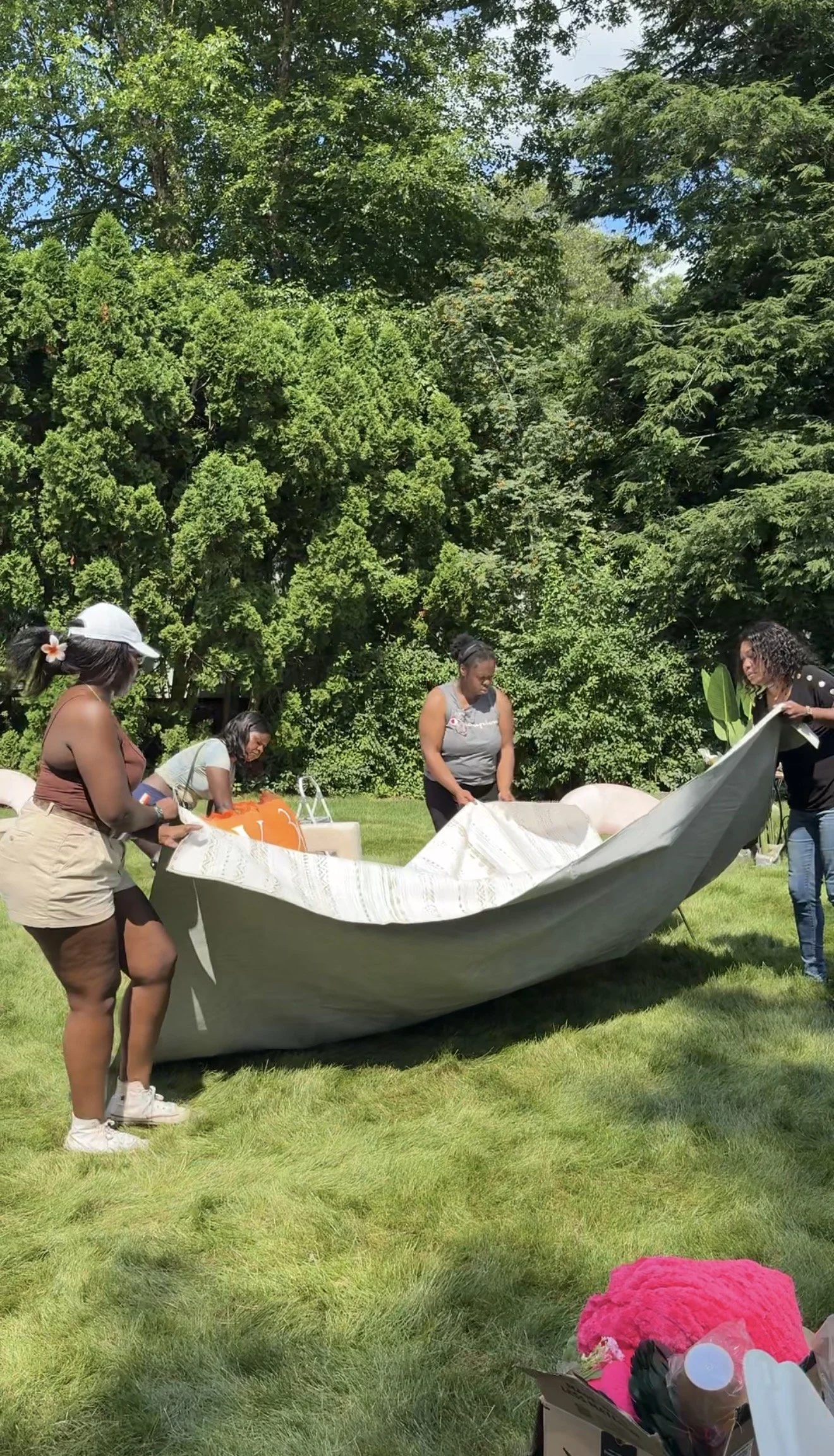 Three women are outside on a grassy area, working together to set up a large tent or canopy. They are surrounded by green trees under a sunny sky. In the foreground, there is a pink fuzzy object and some miscellaneous items.