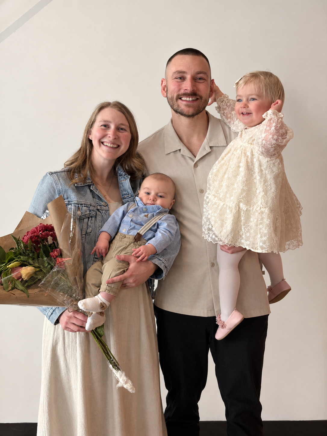 A happy family of four posing together indoors, with the mother holding a bouquet of flowers and the father holding a young girl. The mother is wearing a denim jacket over a beige dress, and the father is wearing a beige shirt with black pants. The young girl is in a lacy cream dress and pink shoes, and a baby boy in a blue shirt and beige pants is being held by the mother.