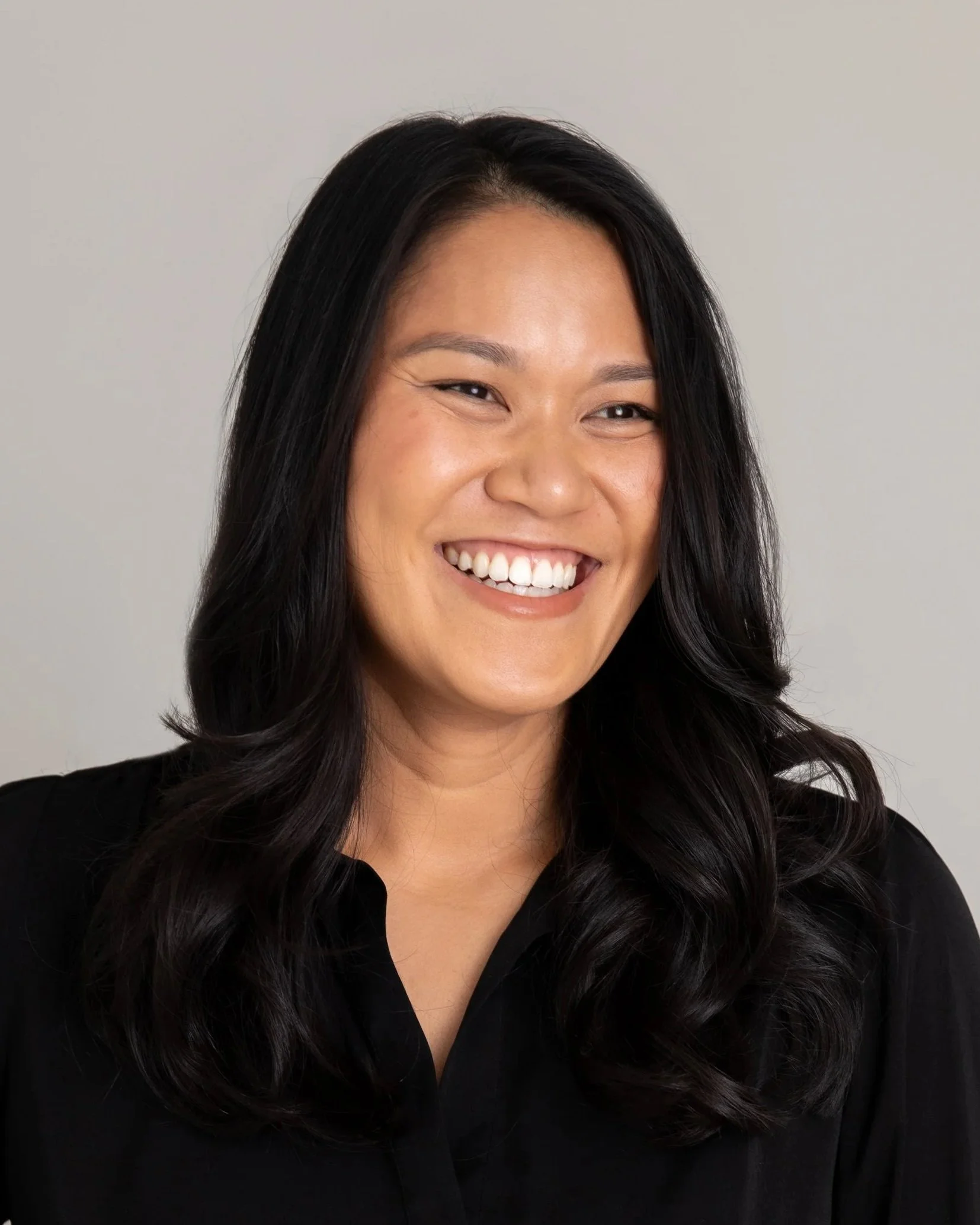 Close-up of a woman with long black hair smiling, wearing a black top, against a plain light background.