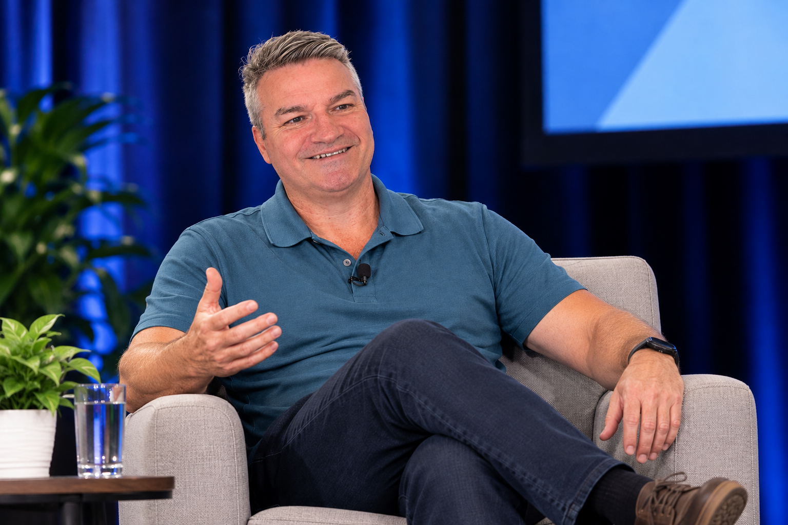 Brett Rutledge in a blue shirt sits on a beige armchair, smiling and gesturing with his right hand during an interview or discussion, with plants and a dark blue curtain in the background.