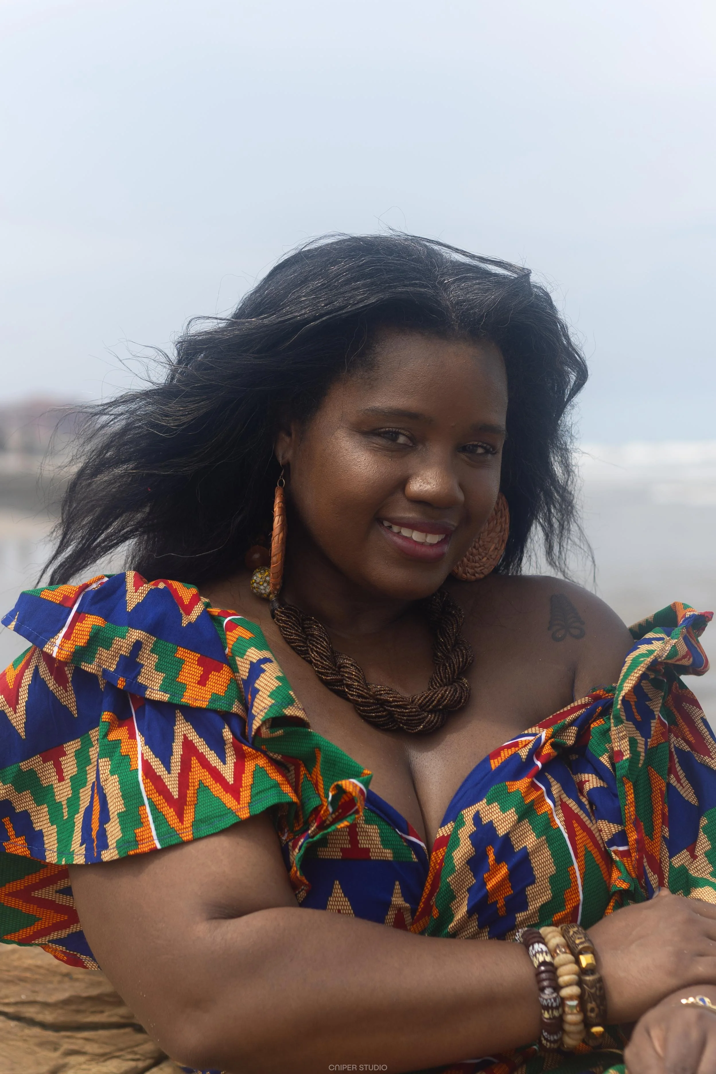Keena wearing a colorful African print dress while seated on rocks along the ocean.