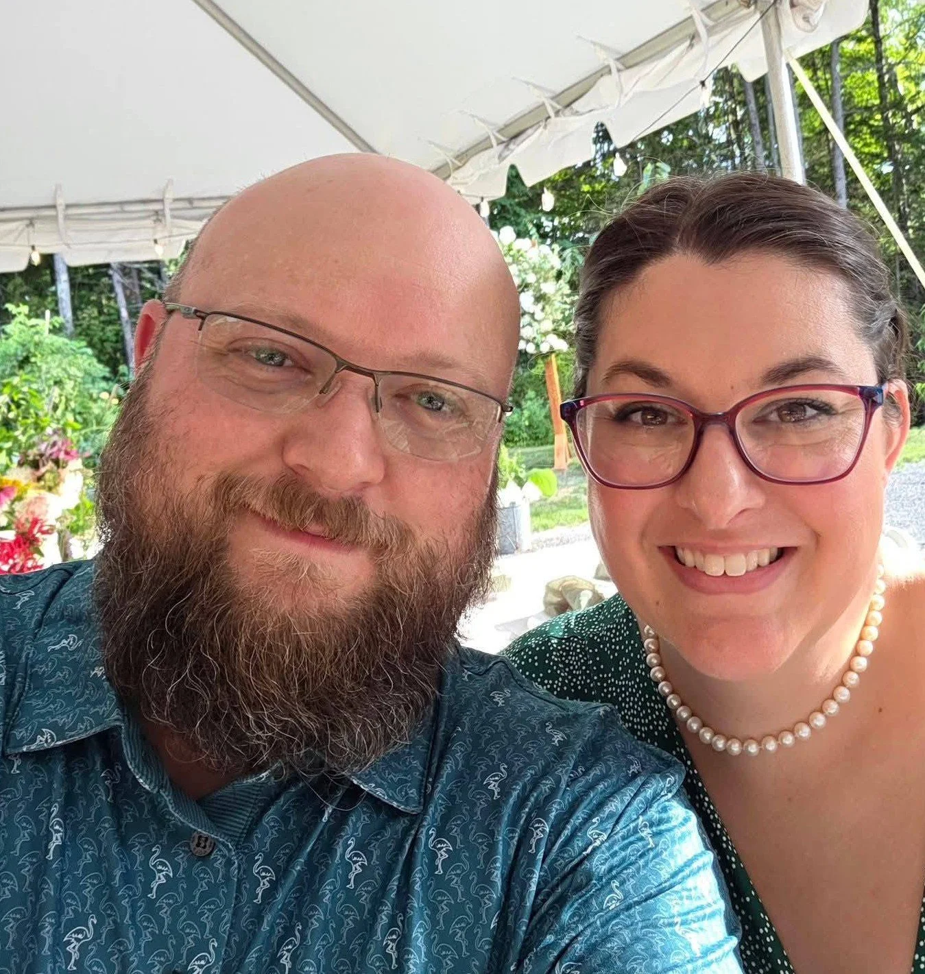 A smiling man with a beard and glasses taking a selfie with a smiling woman with glasses and a pearl necklace outdoors under a tent, with greenery and flowers in the background.