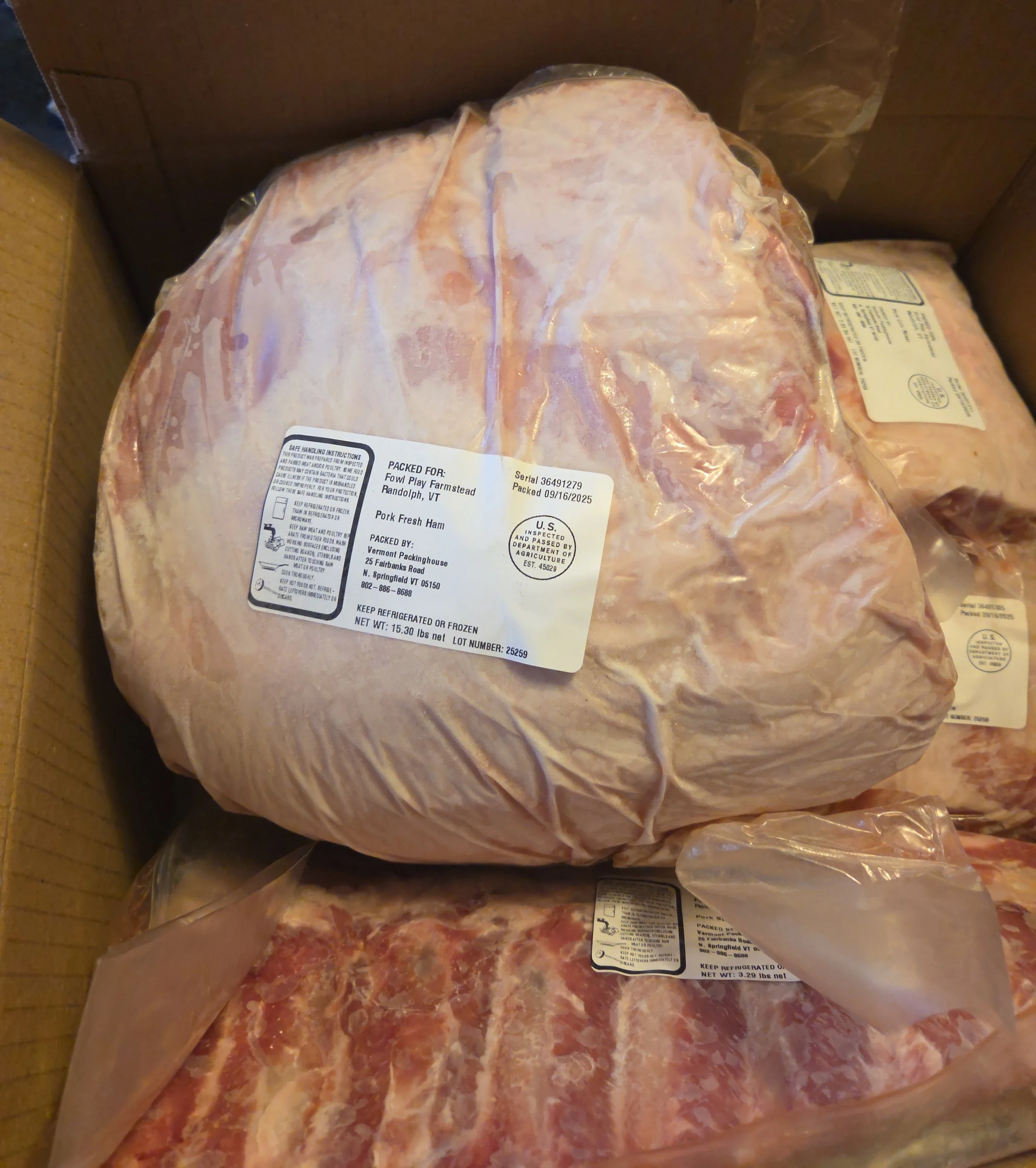 Two packages of raw meat in plastic wrapping inside a cardboard box. The top package contains pork, with a label indicating it is pork ham, while the bottom package appears to contain pig parts with a marbled red and white appearance.