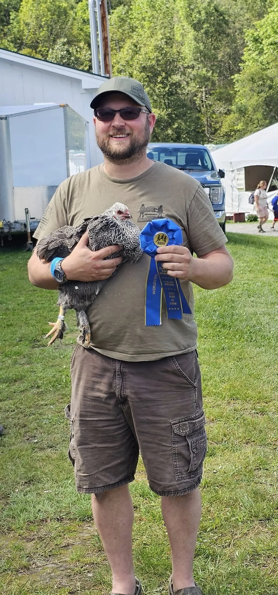 A man in sunglasses, a cap, and casual clothing is holding a turkey and a blue ribbon at an outdoor event with tents and trees in the background.