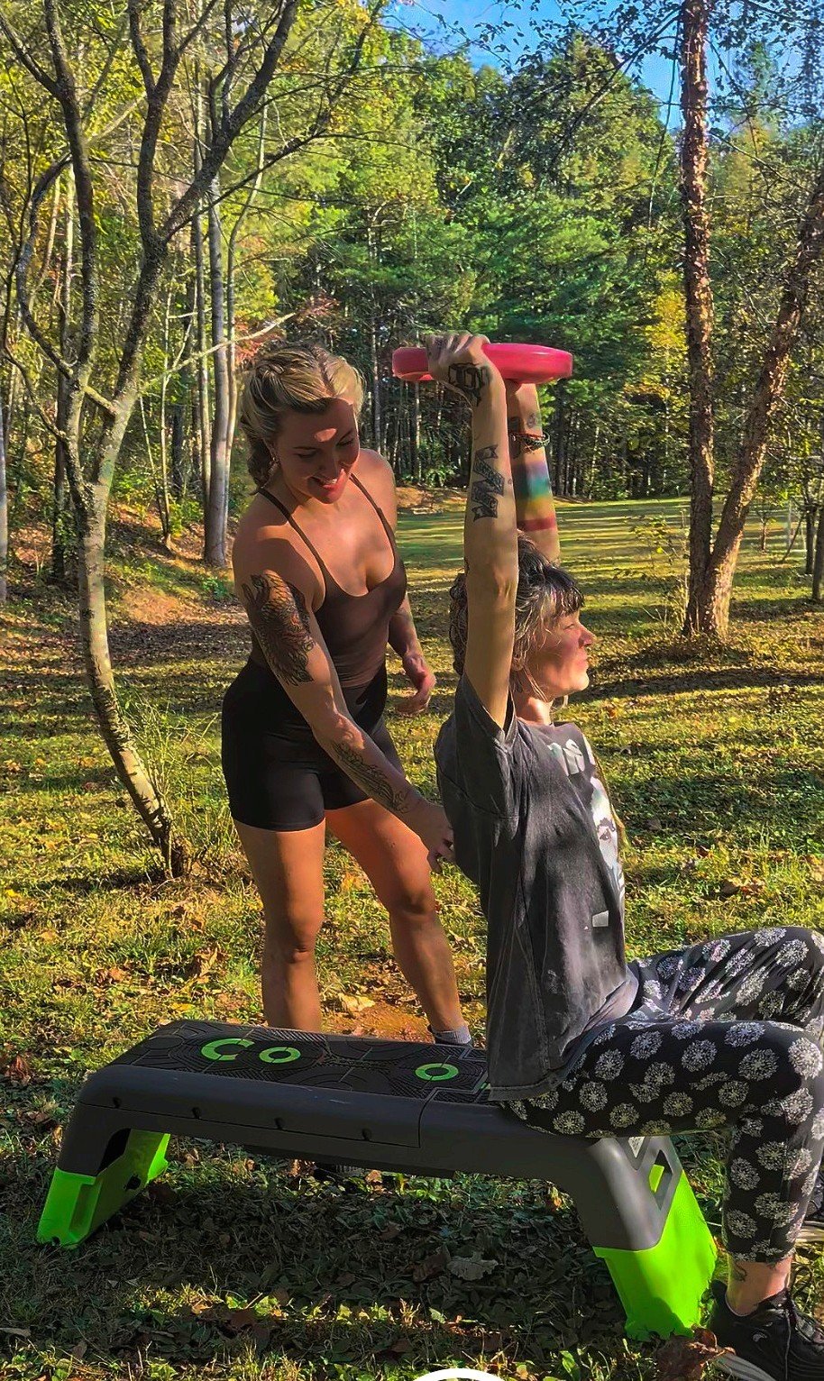 Rachel Huncke has tattoos and is assisting another woman with tattoos, sitting on a bench outdoors in a wooded area, lifting a pink weight overhead.