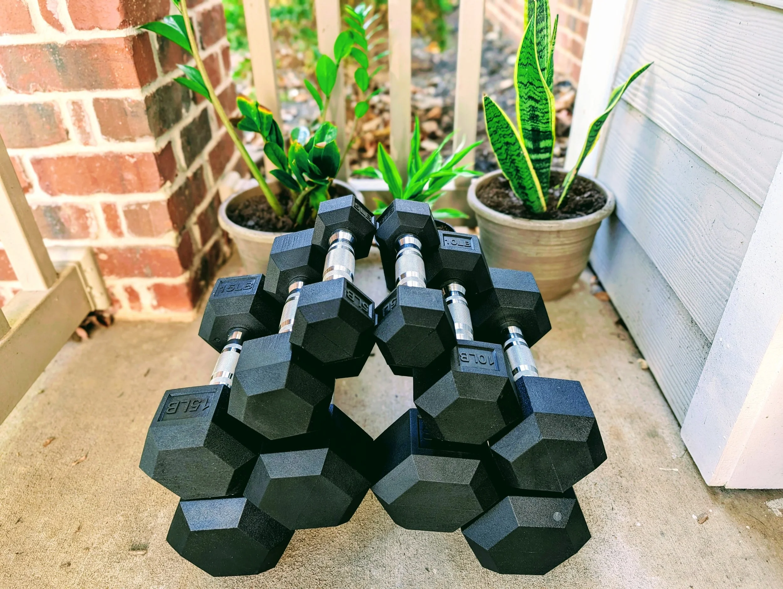 A stack of black hexagonal dumbbells with silver handles placed on a concrete porch, with potted plants and a brick and white siding wall in the background.