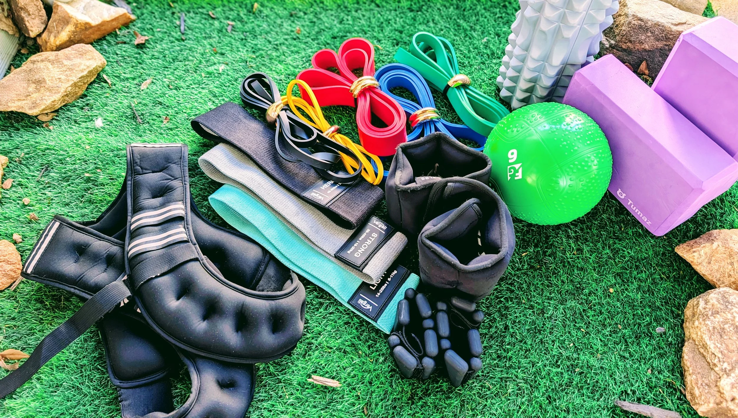 An array of fitness and sports equipment including resistance bands, a kettlebell, a medicine ball, a foam roller, ankle weights, and other accessories are displayed on artificial green grass outdoors.