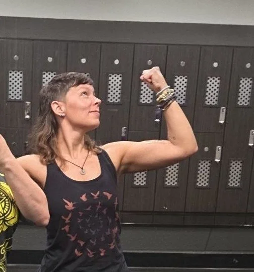 A woman in a gym locker room flexing her right bicep and looking at it, wearing a black tank top with a bird graphic and various bracelets.
