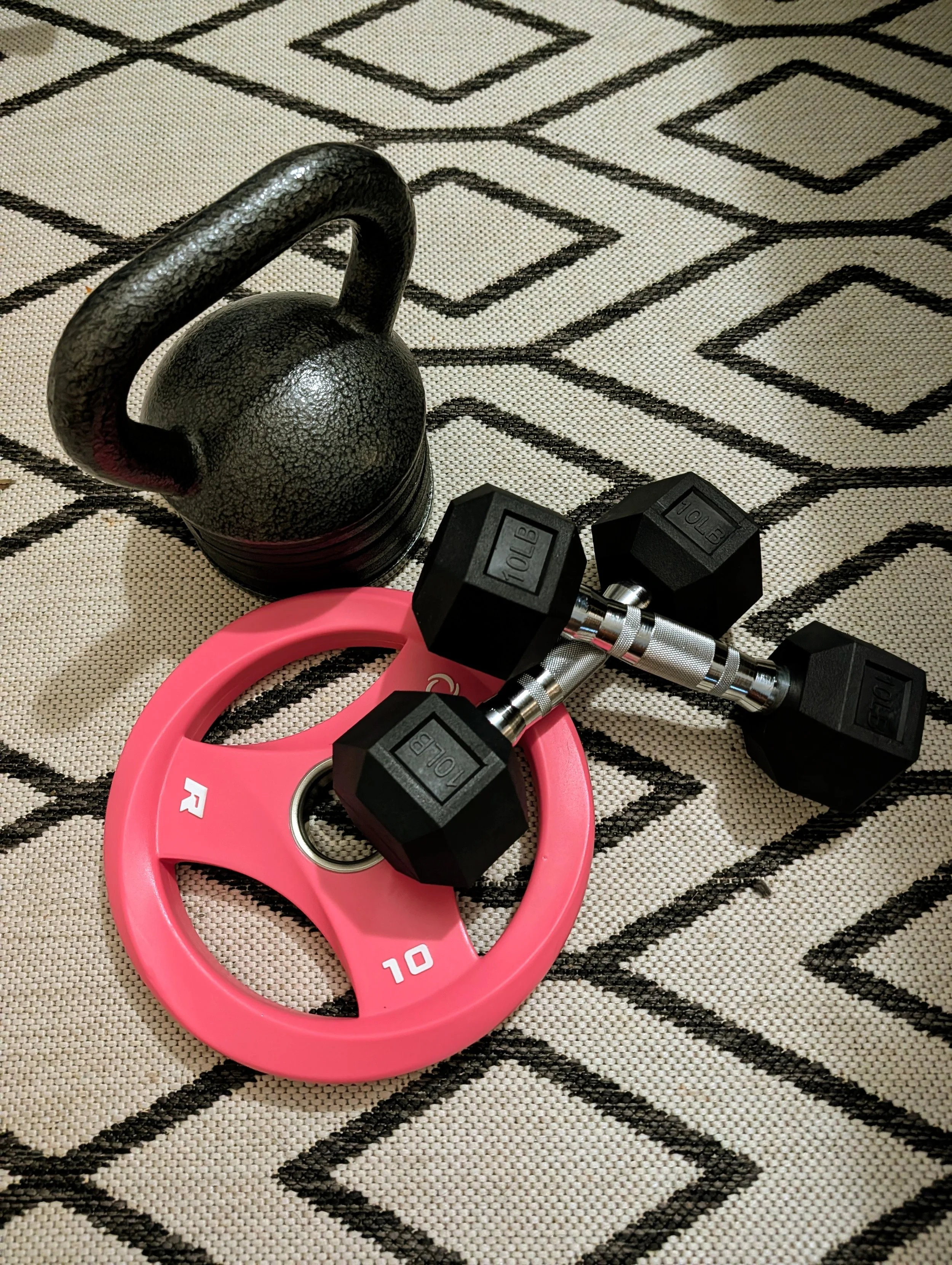 A black kettlebell, two small black dumbbells, a pink weight plate, and a pink circular resistance band are placed on a black and white patterned mat.