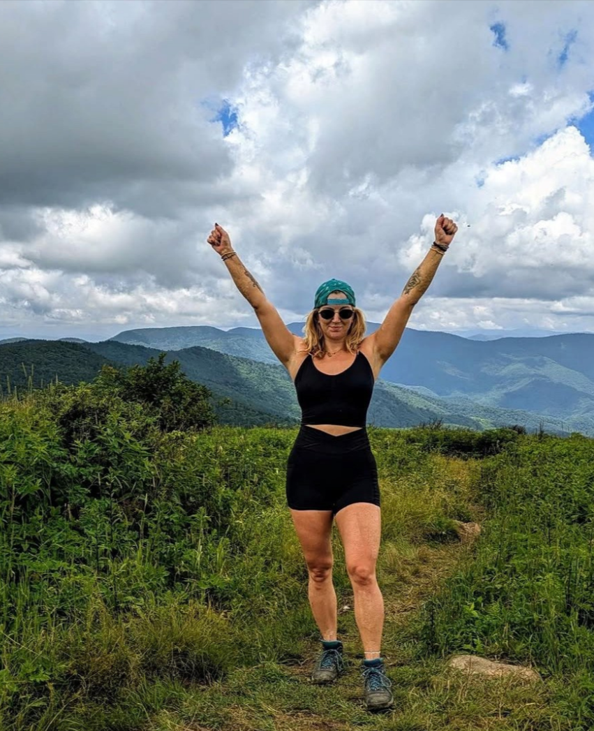 Rachel Huncke standing on a hiking trail in a green mountainous area, raising her arms in triumph, wearing a black sports bra, black shorts, sunglasses, sneakers, and a blue cap, with a cloudy sky overhead.
