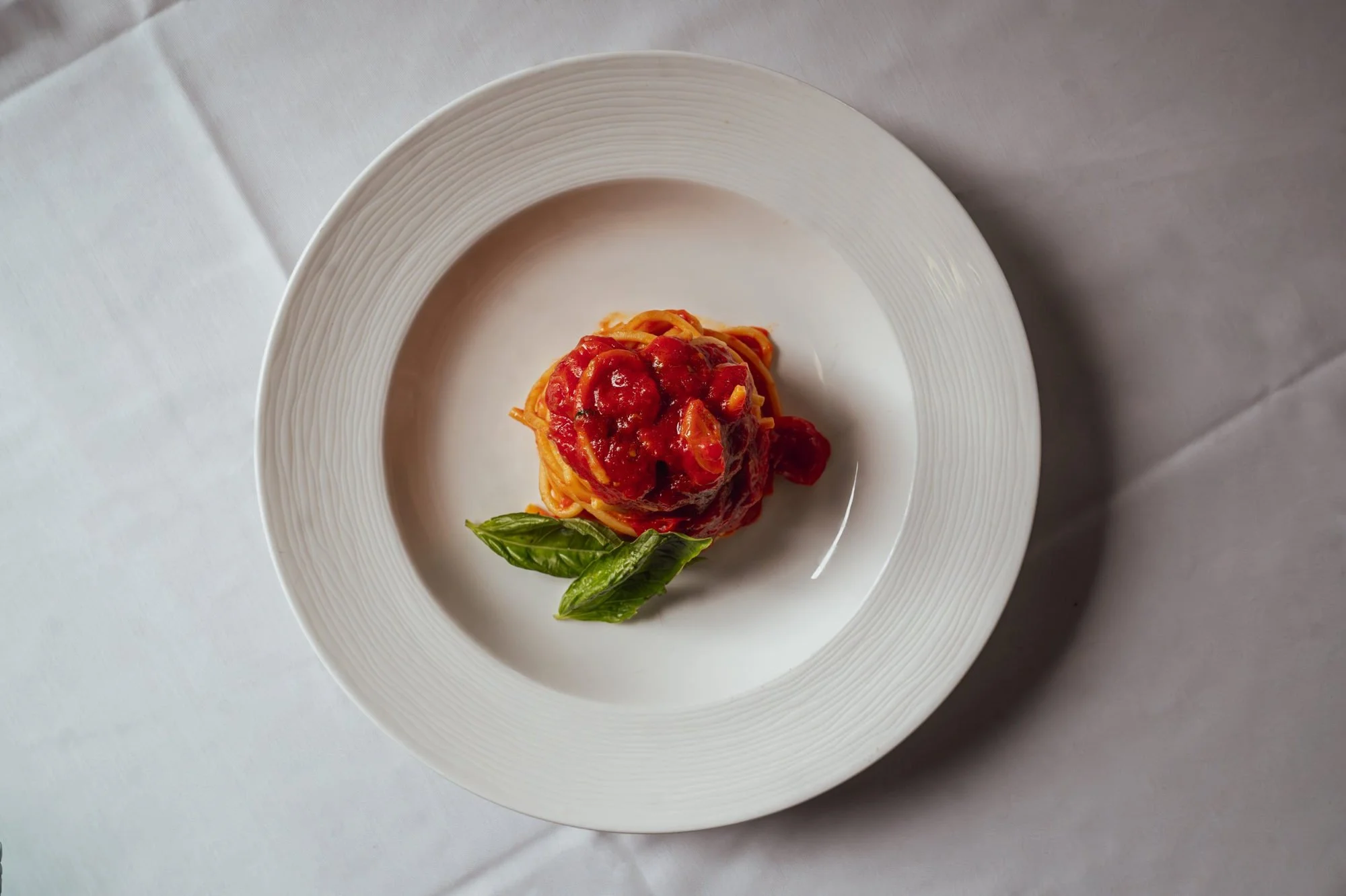 A plate of spaghetti with tomato sauce and fresh basil leaves served on a white textured plate.
