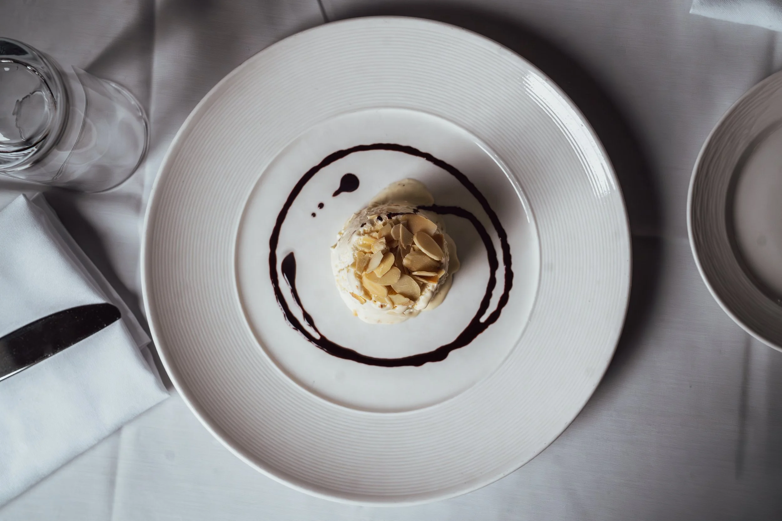 Dessert with caramel and almond slices on a white plate, decorated with chocolate syrup, placed on a metal surface with glasses and napkins nearby.