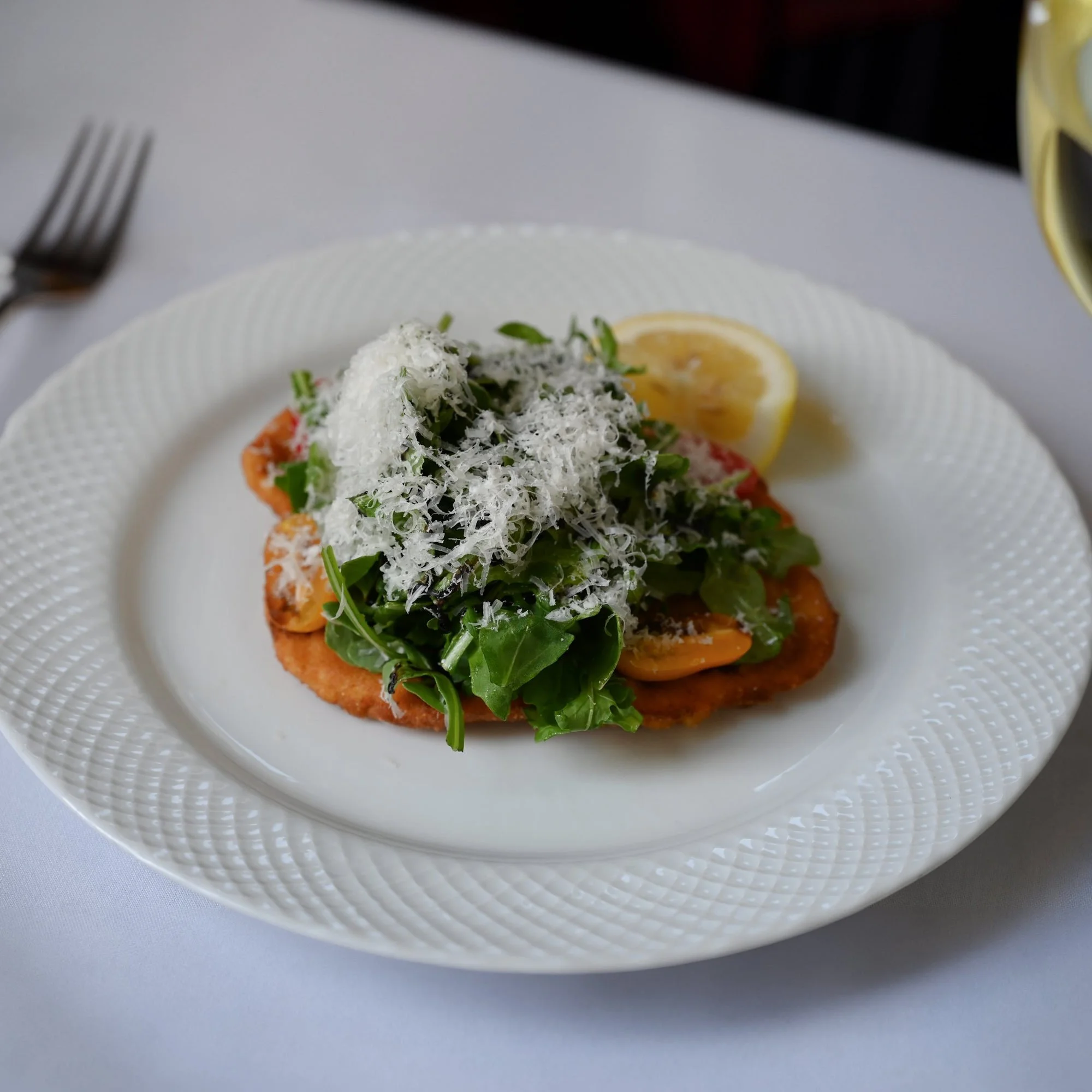 Plate with food including a lemon wedge, shredded white cheese, chopped herbs, and a slice of tomato, on a white textured plate.