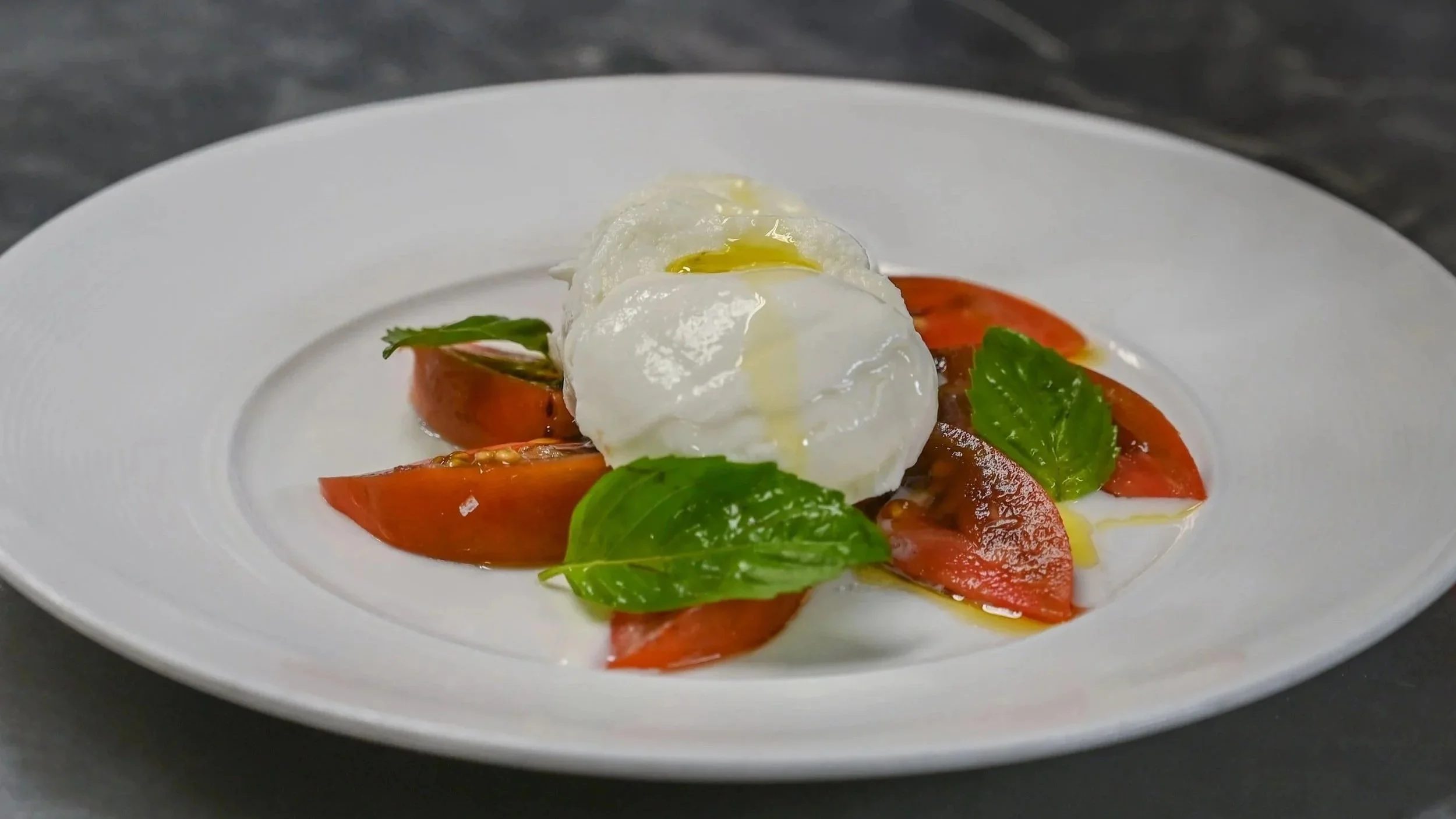 A plate of sliced tomatoes topped with fresh basil leaves, with three poached eggs drizzled with olive oil in the center.