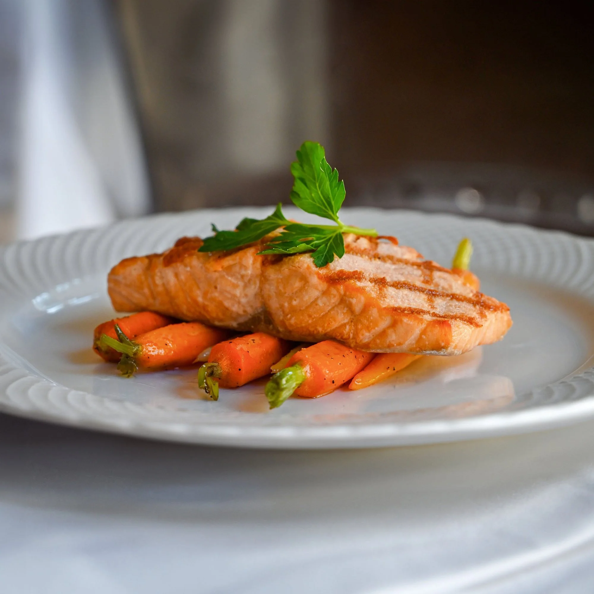 Close-up of a plated dish featuring grilled or baked salmon garnished with a fresh green parsley leaf, served with a side of grilled carrots, viewed through a glass oven door.