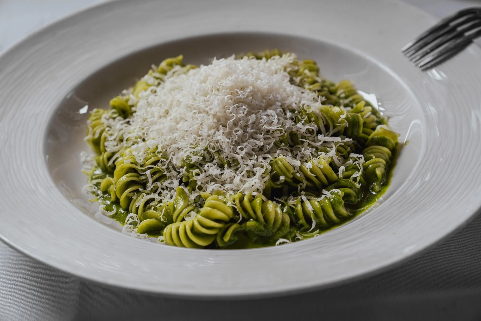 Plate of green pesto fusilli pasta topped with grated cheese, fork and knife on the side.