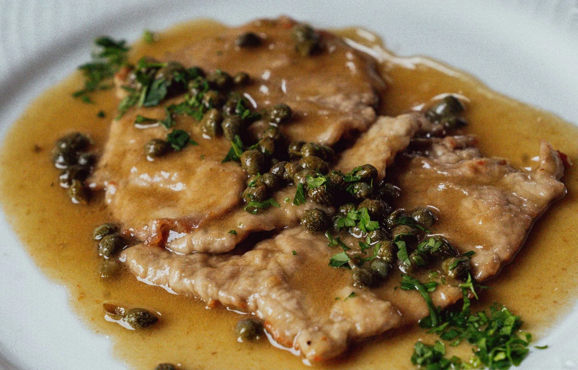 A plated dish featuring a piece of cooked fish topped with capers and chopped herbs, served with a light sauce or broth.